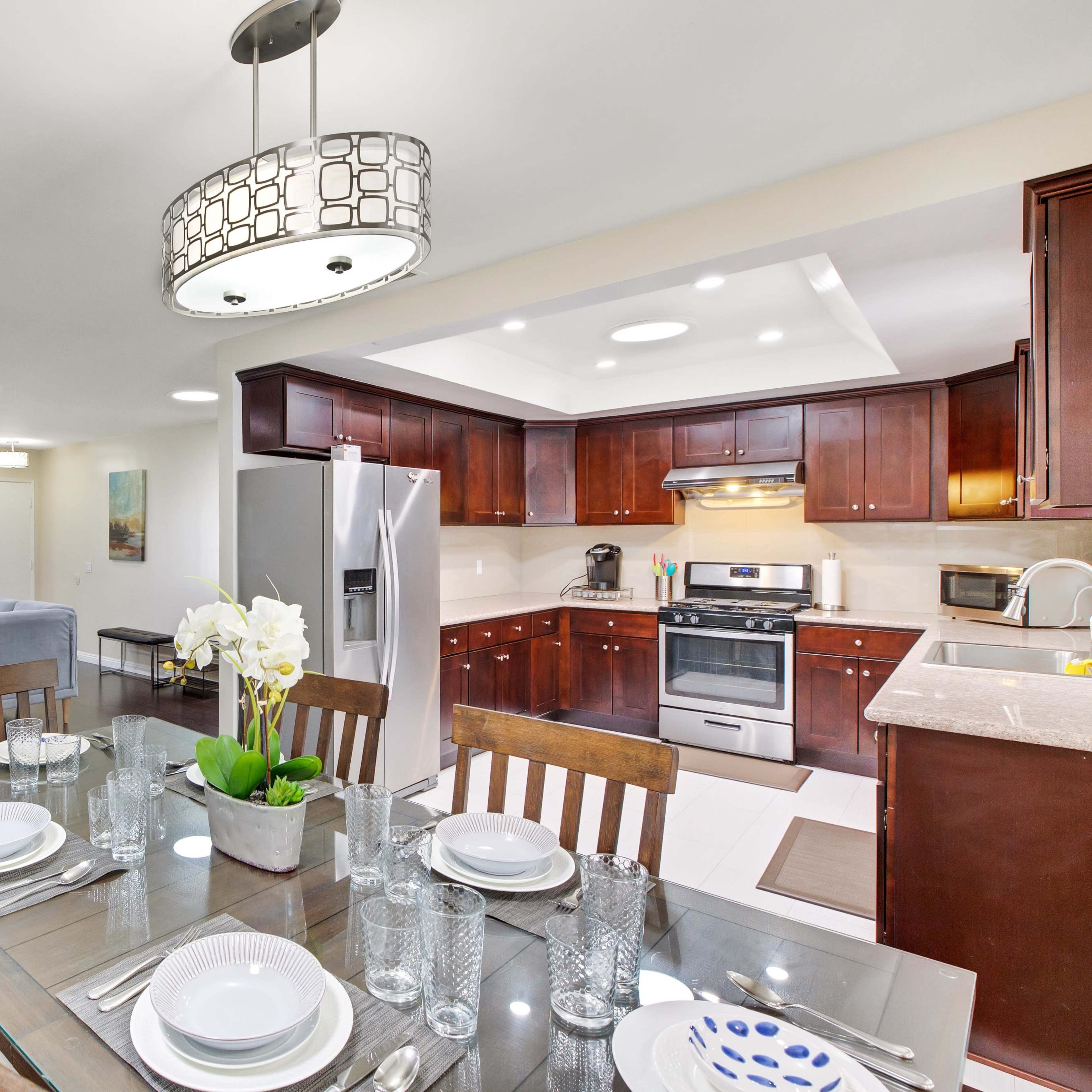 A modern kitchen and dining area features dark wooden cabinets, stainless steel appliances, and a glass dining table with place settings.