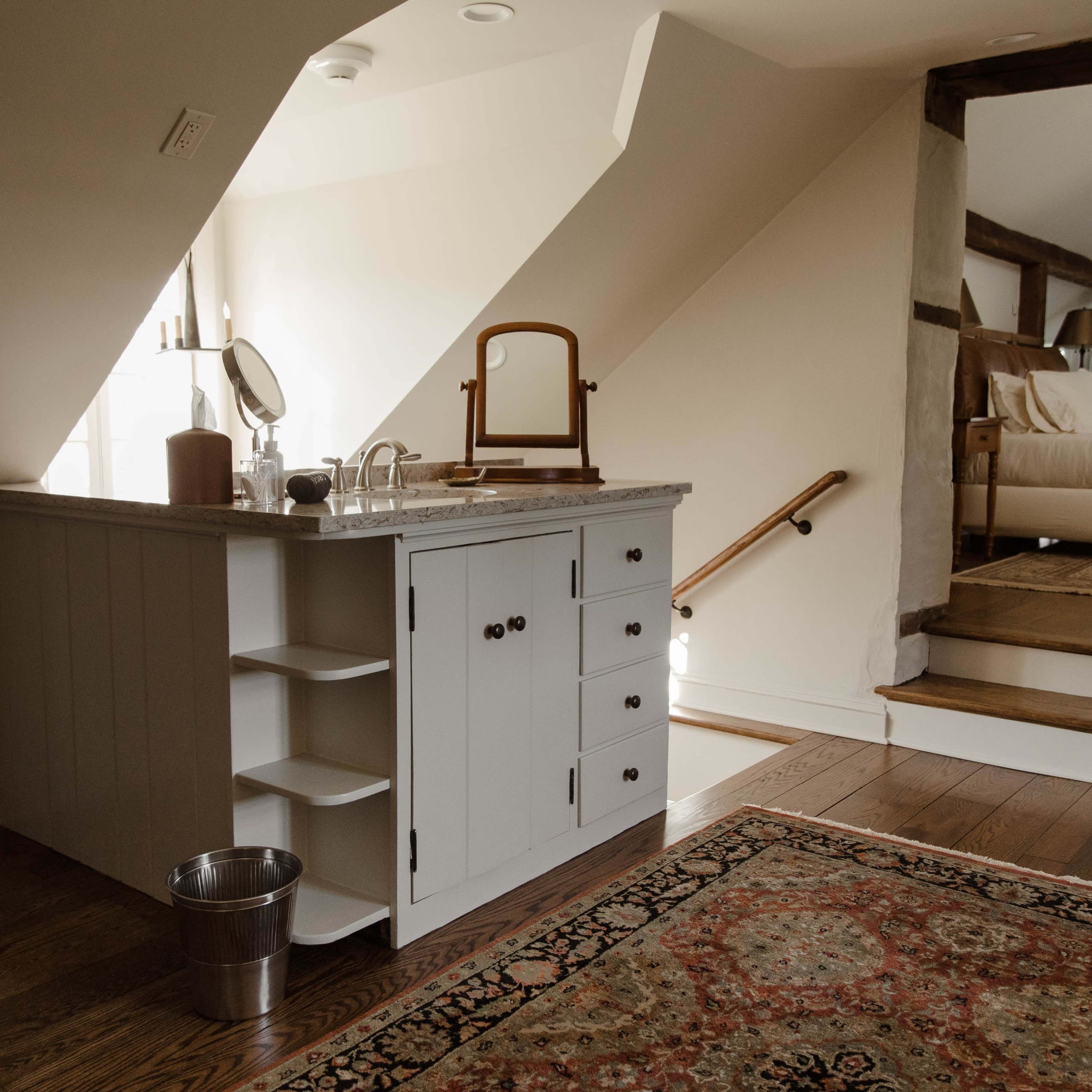 A cozy attic space featuring a small counter with storage drawers, a mirror, and a wastebasket, alongside stairs leading to a bedroom.