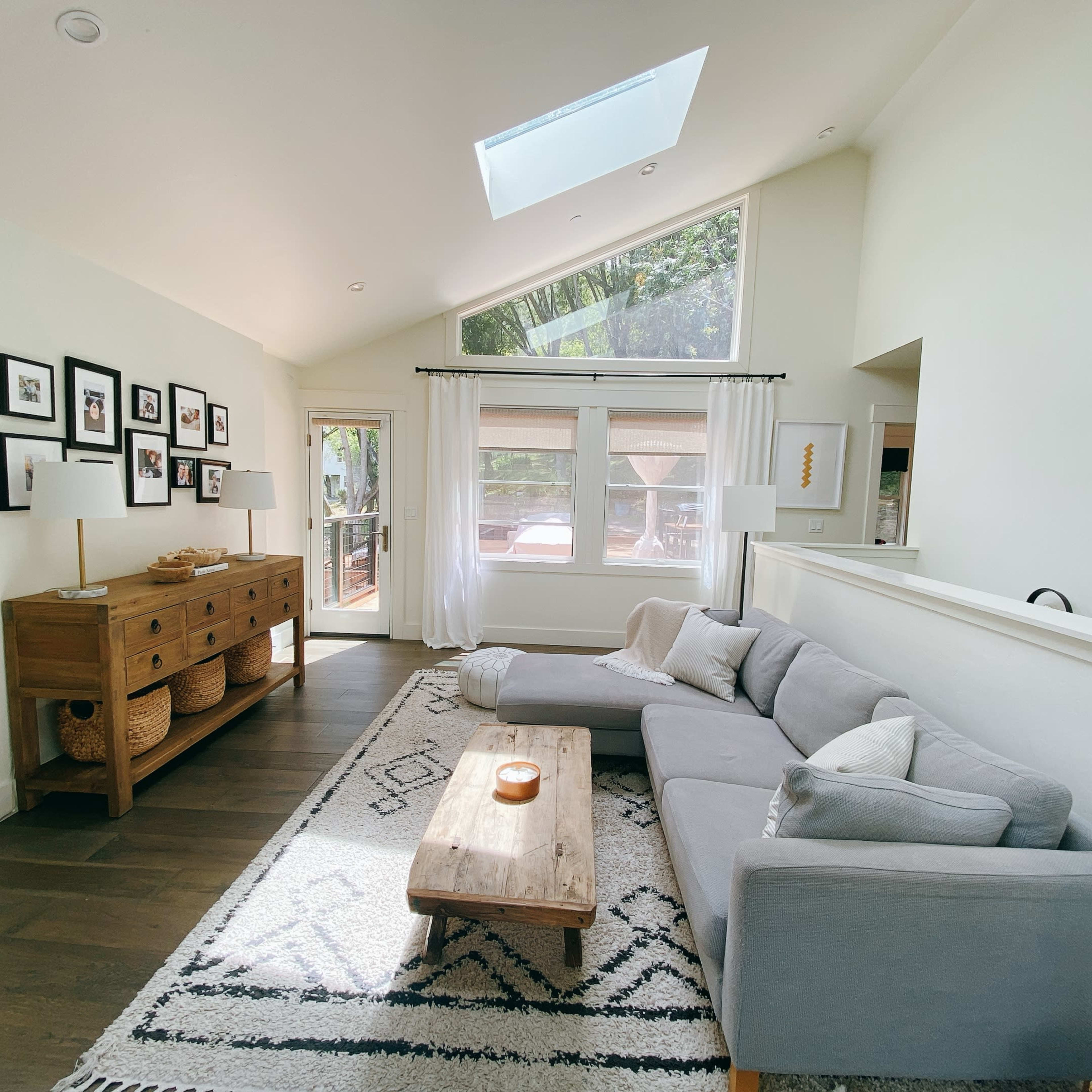 A bright living room featuring a gray sectional sofa, a wooden coffee table, and a console table under framed photographs along the wall.