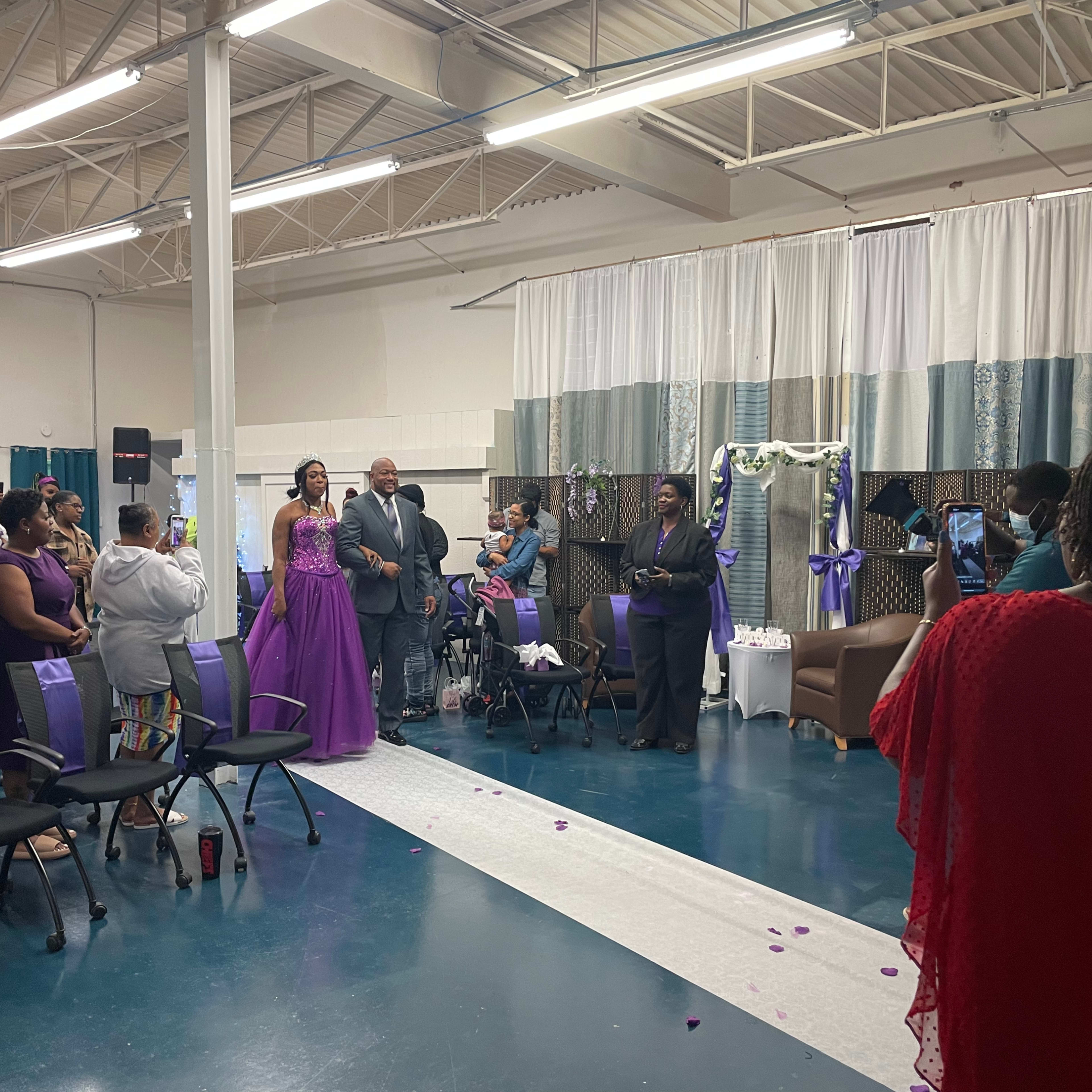 A wedding ceremony is taking place indoors, with guests seated and the bride and groom standing at the front near a decorated backdrop.