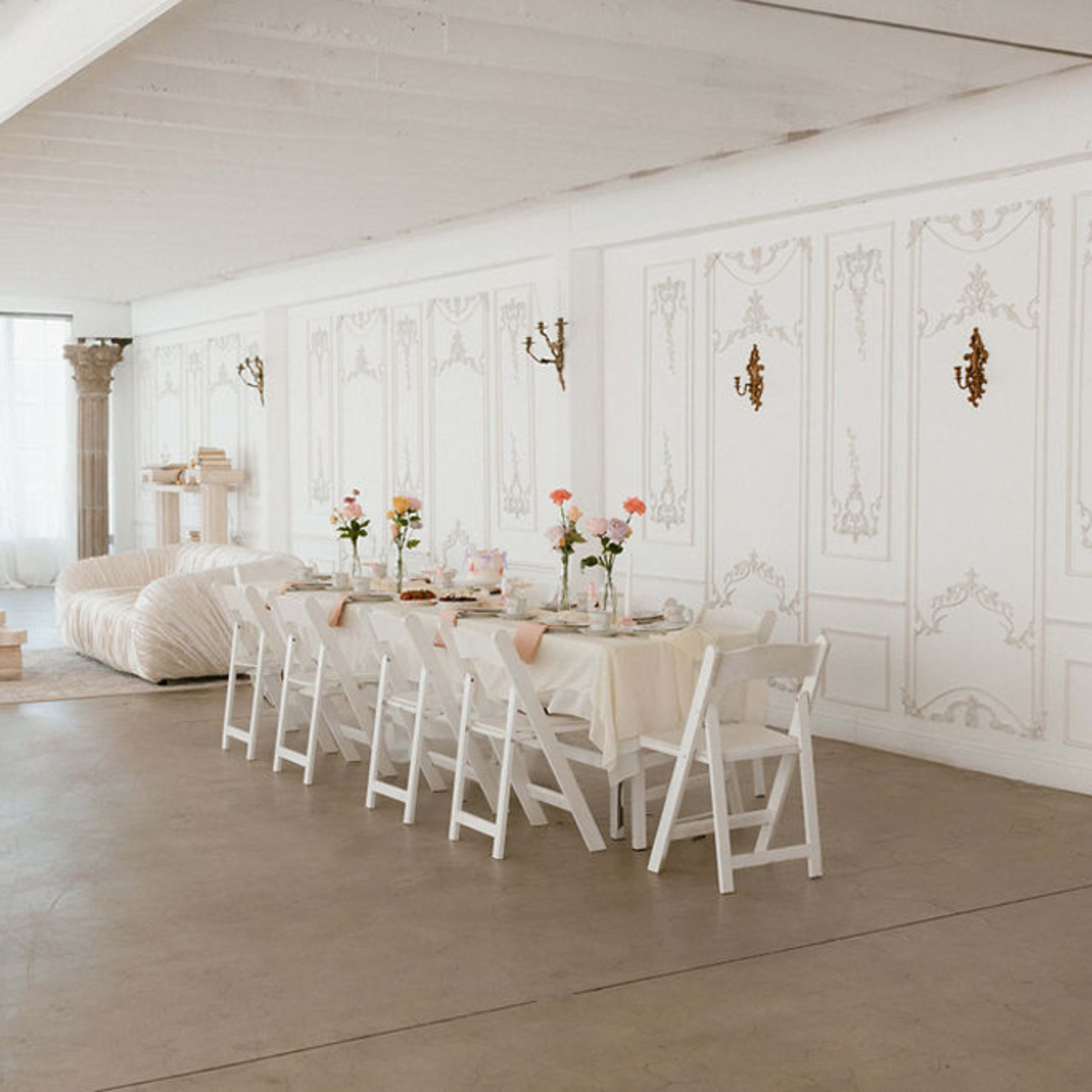 A long, elegantly set dining table with white chairs is positioned in a bright, minimalistic room featuring decorative wall moldings and soft natural light.