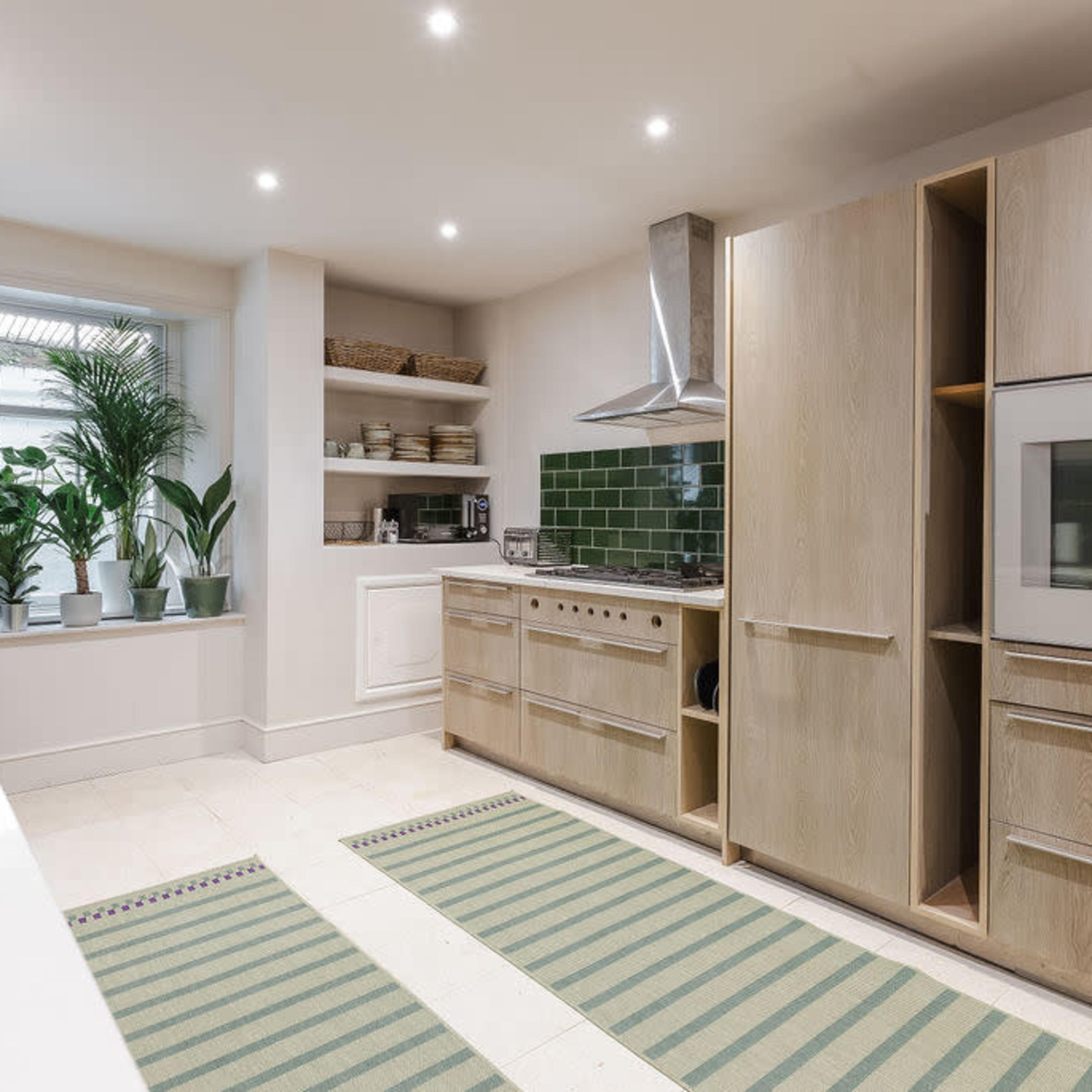 The kitchen features light wood cabinetry, a green tiled backsplash, and a window lined with potted plants.