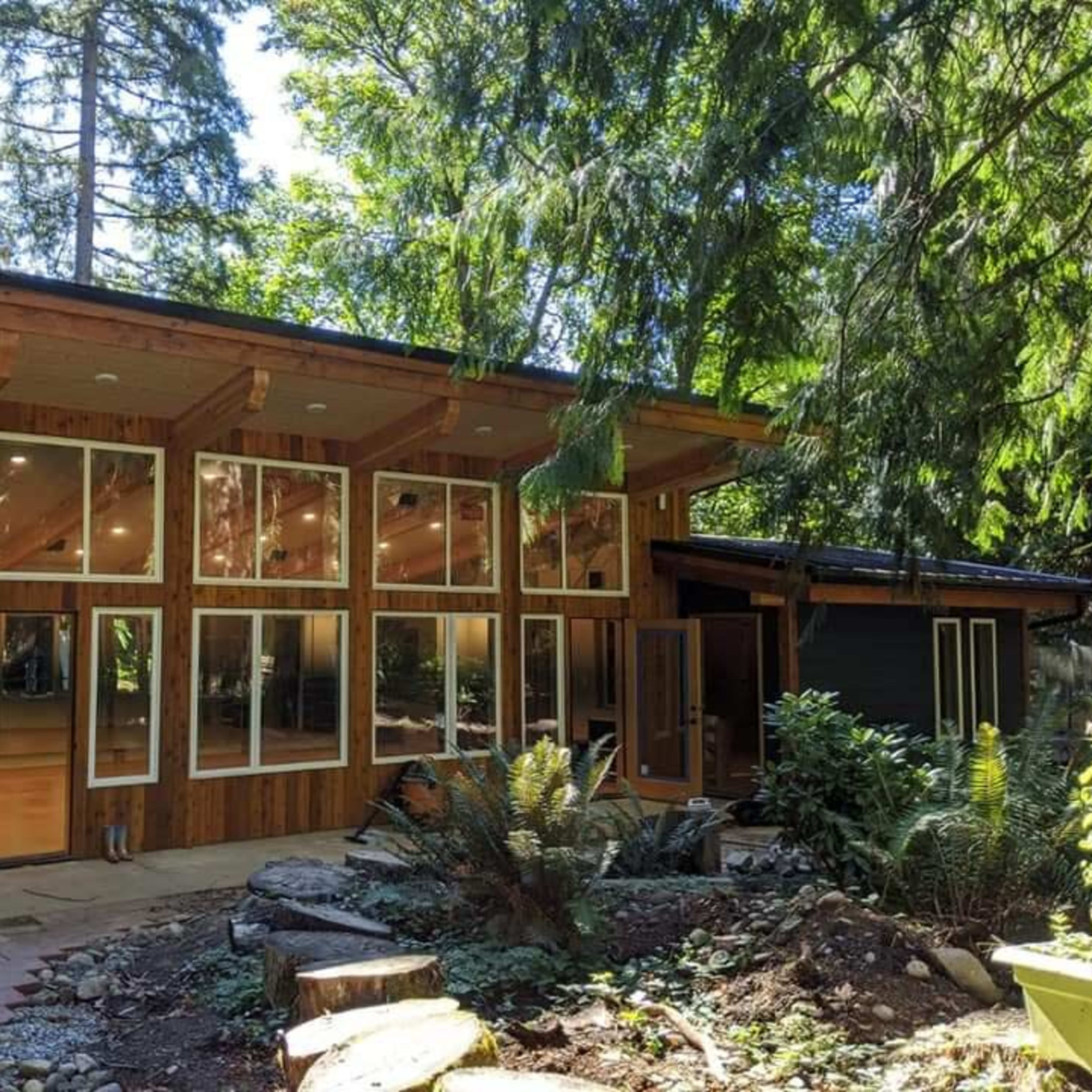 A modern wooden cabin surrounded by trees, featuring large windows and a stone pathway leading to the entrance.