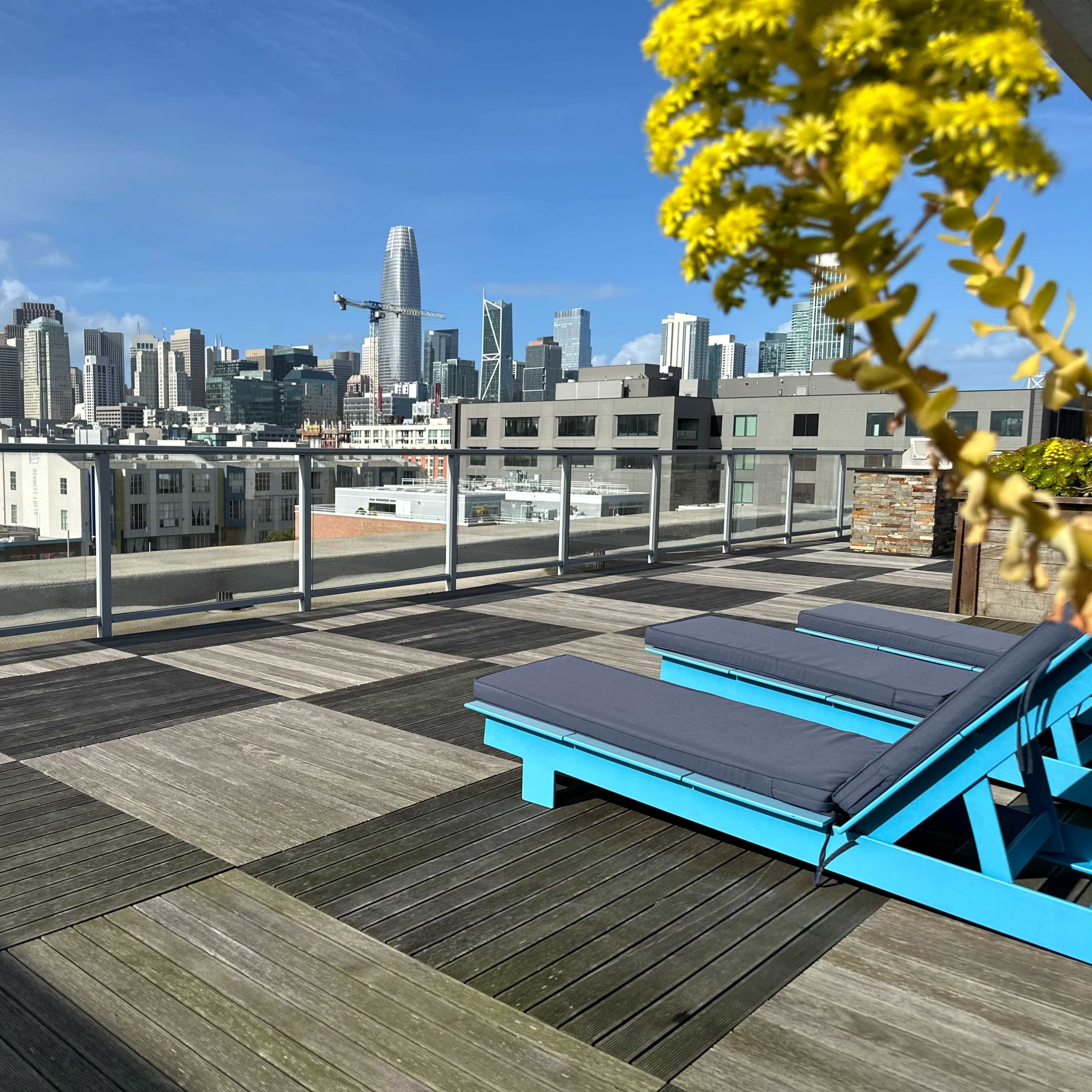 A rooftop terrace with blue lounge chairs overlooks a skyline filled with tall buildings under a clear blue sky.