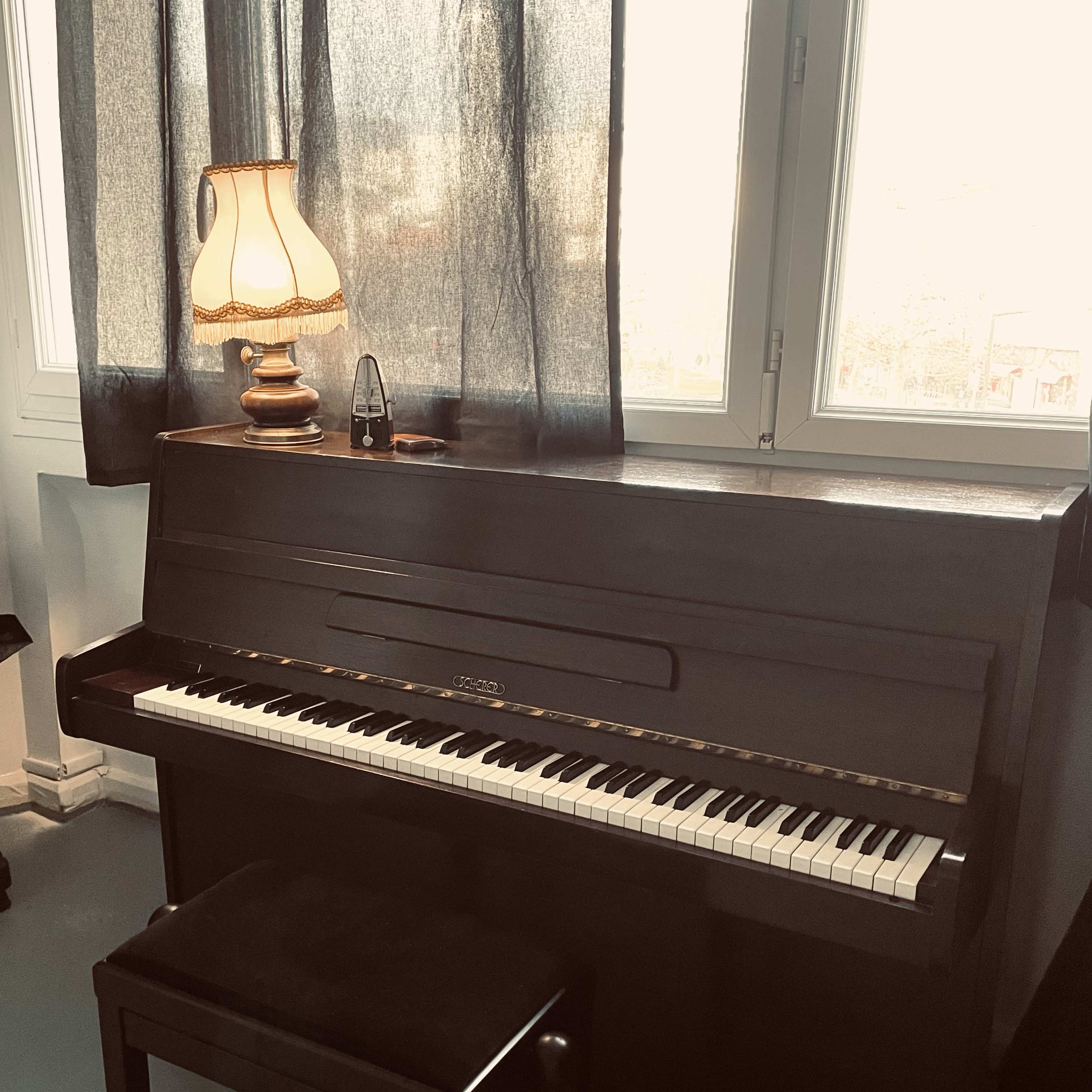 A black piano with a matching bench is positioned next to a window, accompanied by a lamp on top.