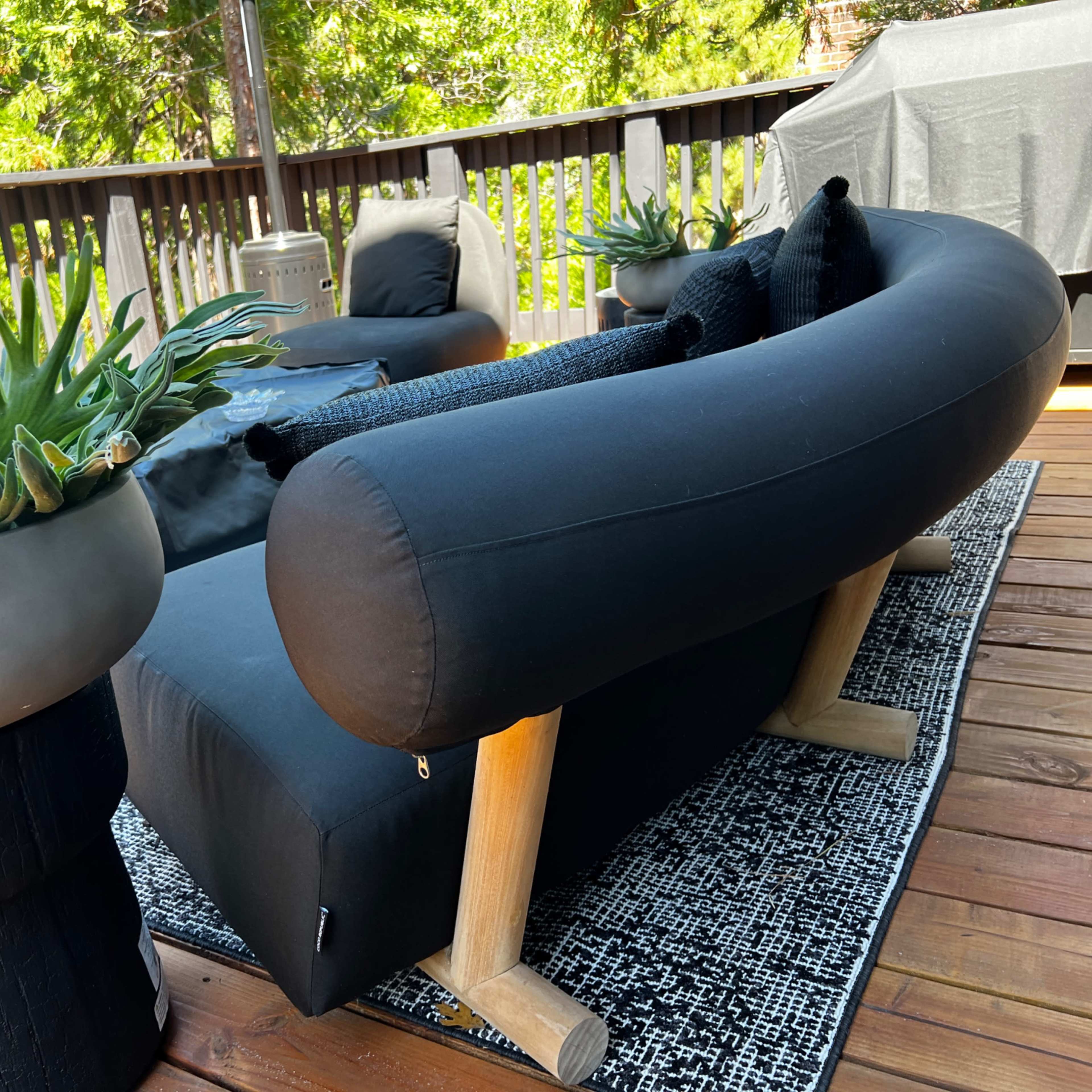 A modern outdoor seating arrangement with a curved black sofa and a large planter filled with plants on a wooden deck.