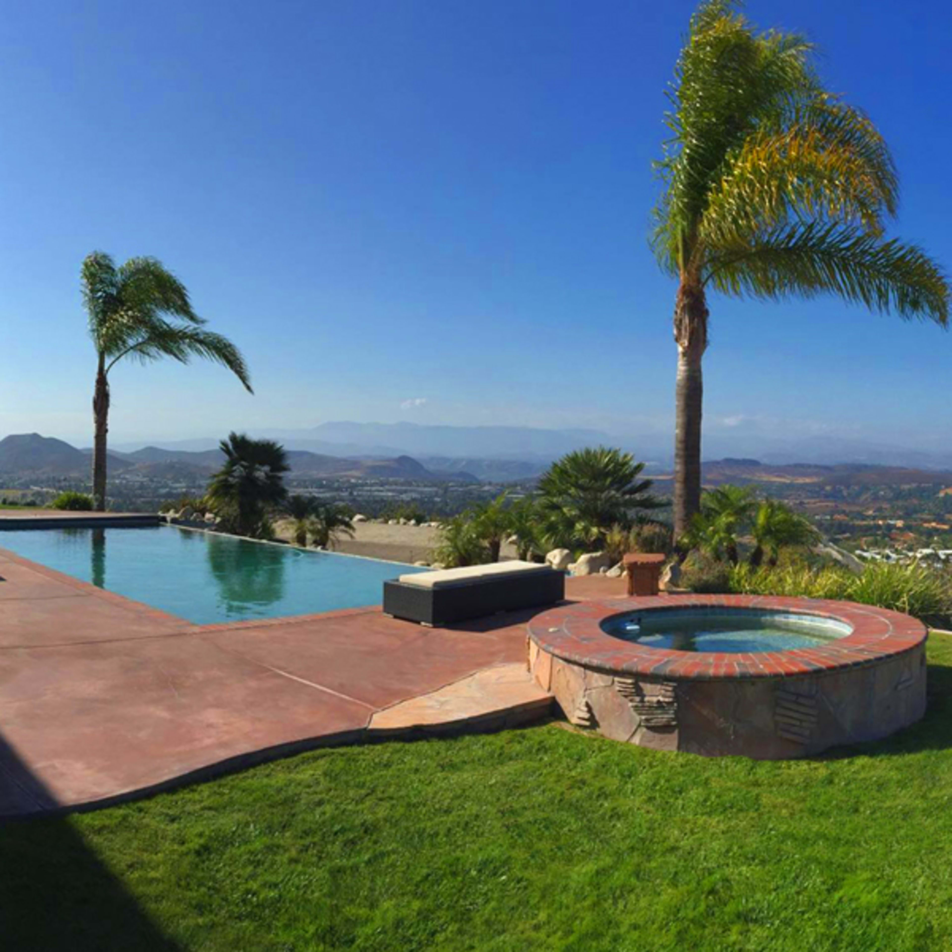 A swimming pool and hot tub surrounded by a grassy area with palm trees, overlooking a valley and distant mountains under a clear blue sky.