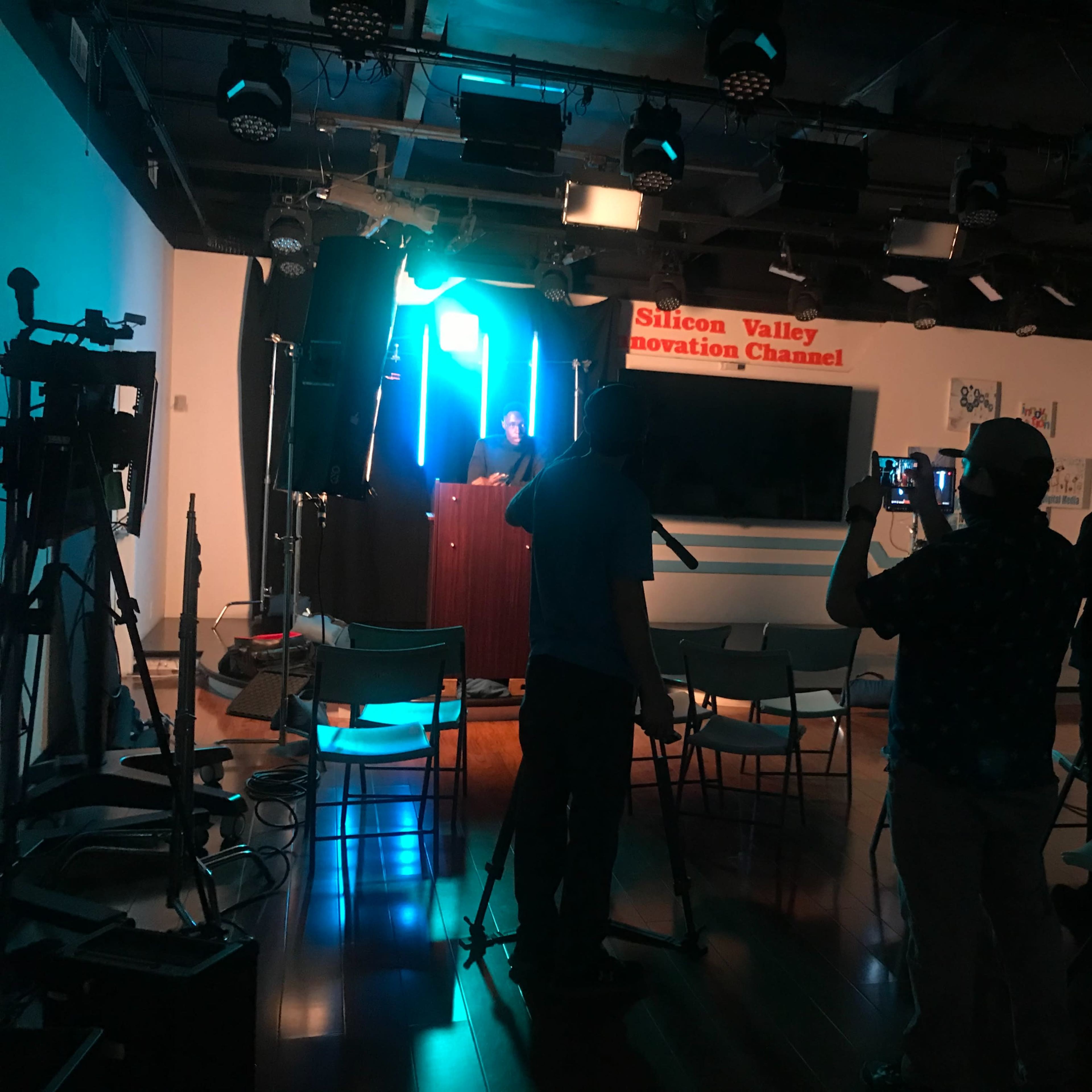 A person stands at a podium in a studio setting, while cameras and a small audience are positioned in front of them.