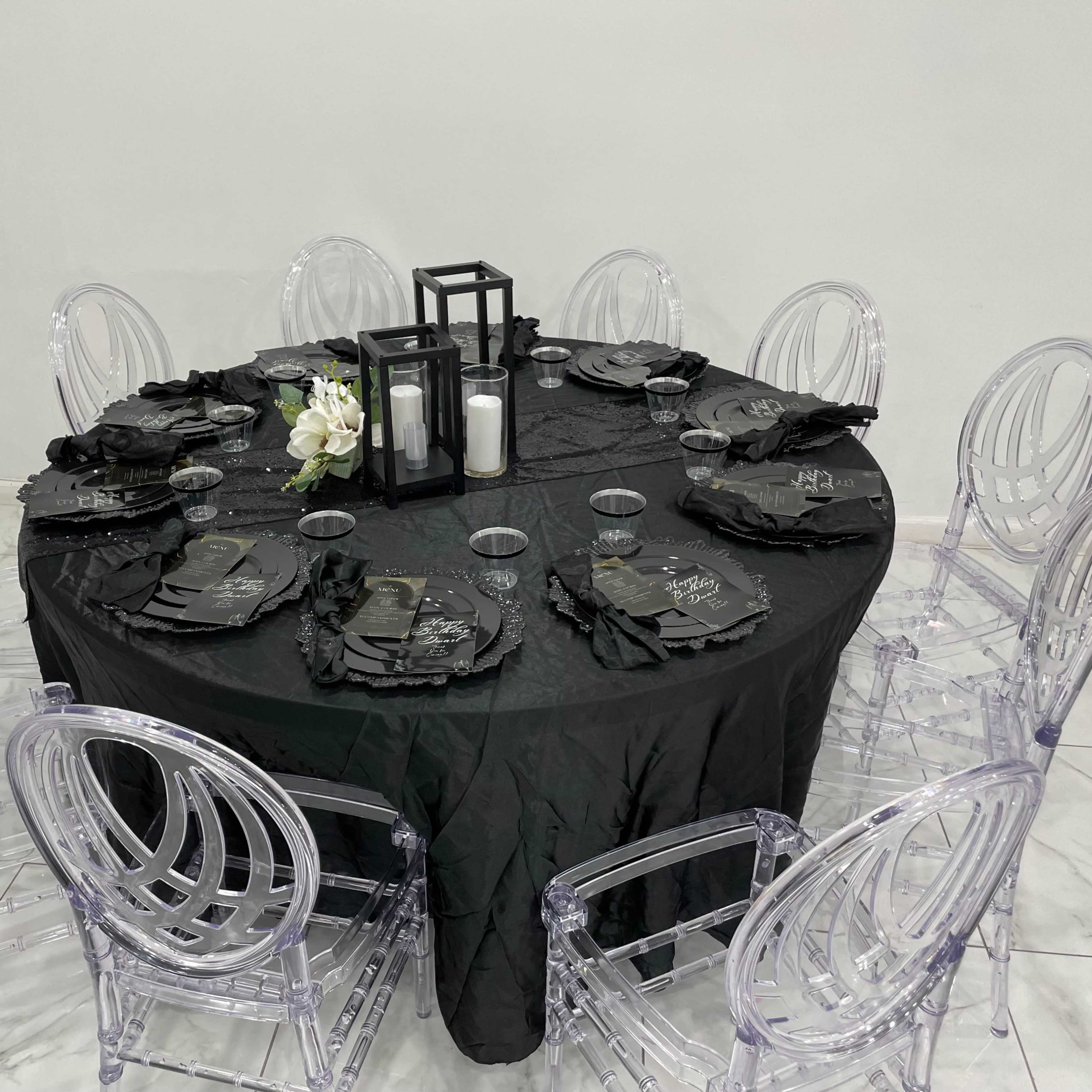 A round table is set with a black tablecloth, clear chairs, black plates, and decorative candle holders, surrounded by twelve chairs.