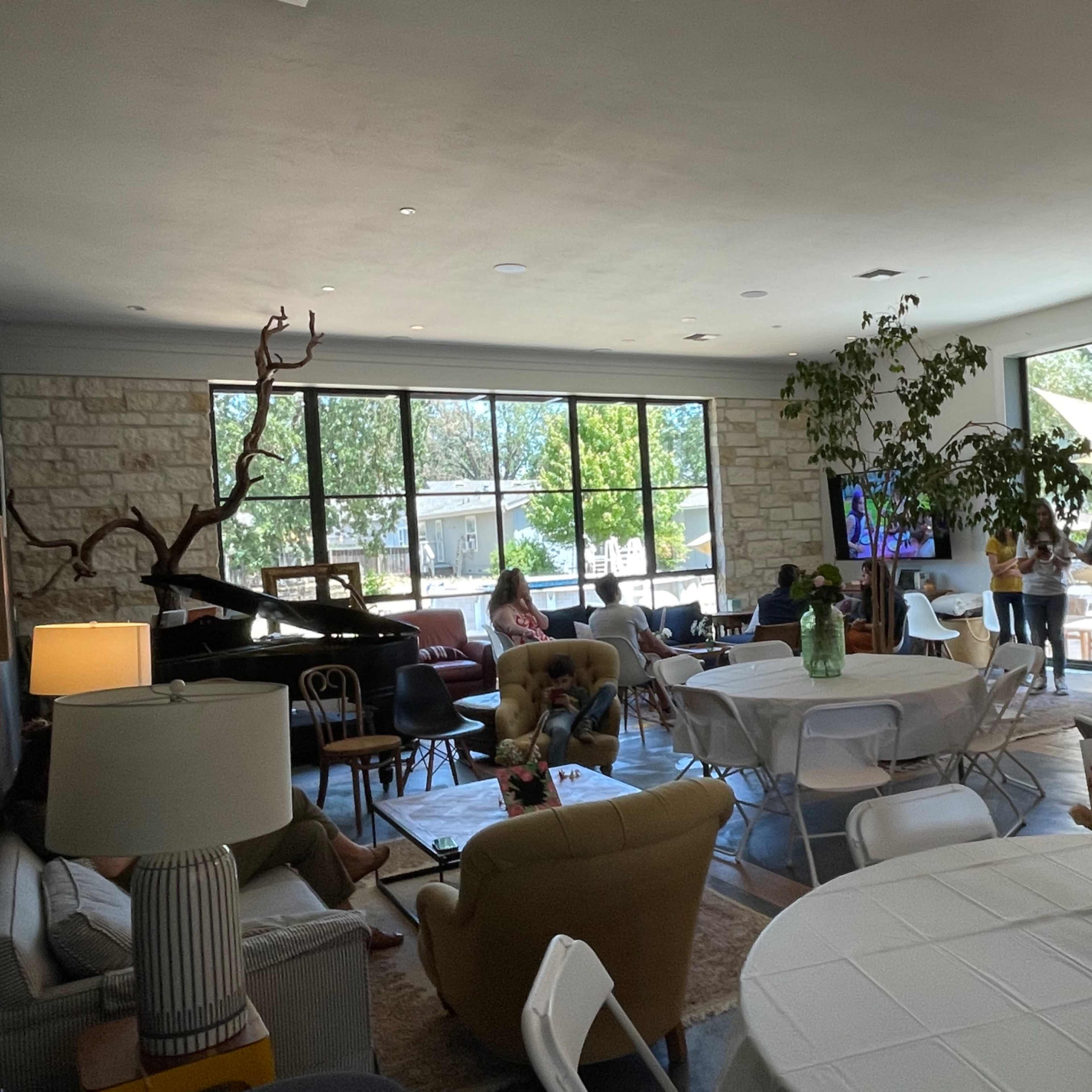 A spacious room with large windows, featuring a mix of seating areas, tables set for gatherings, and a piano in one corner.