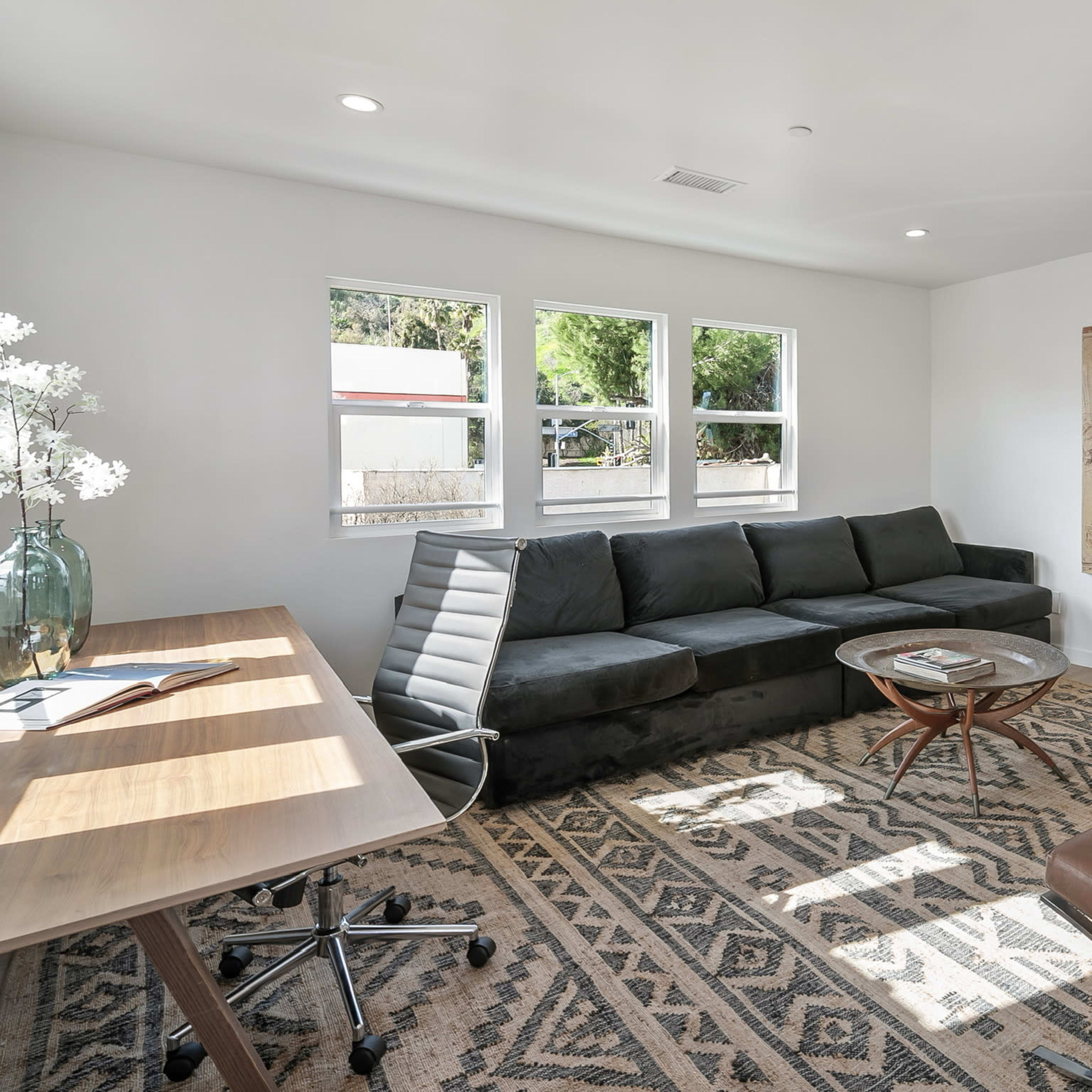 A modern living area with a dark sectional sofa, a glass coffee table, a wooden desk with an office chair, and two leather lounge chairs, all set against a neutral-colored wall.
