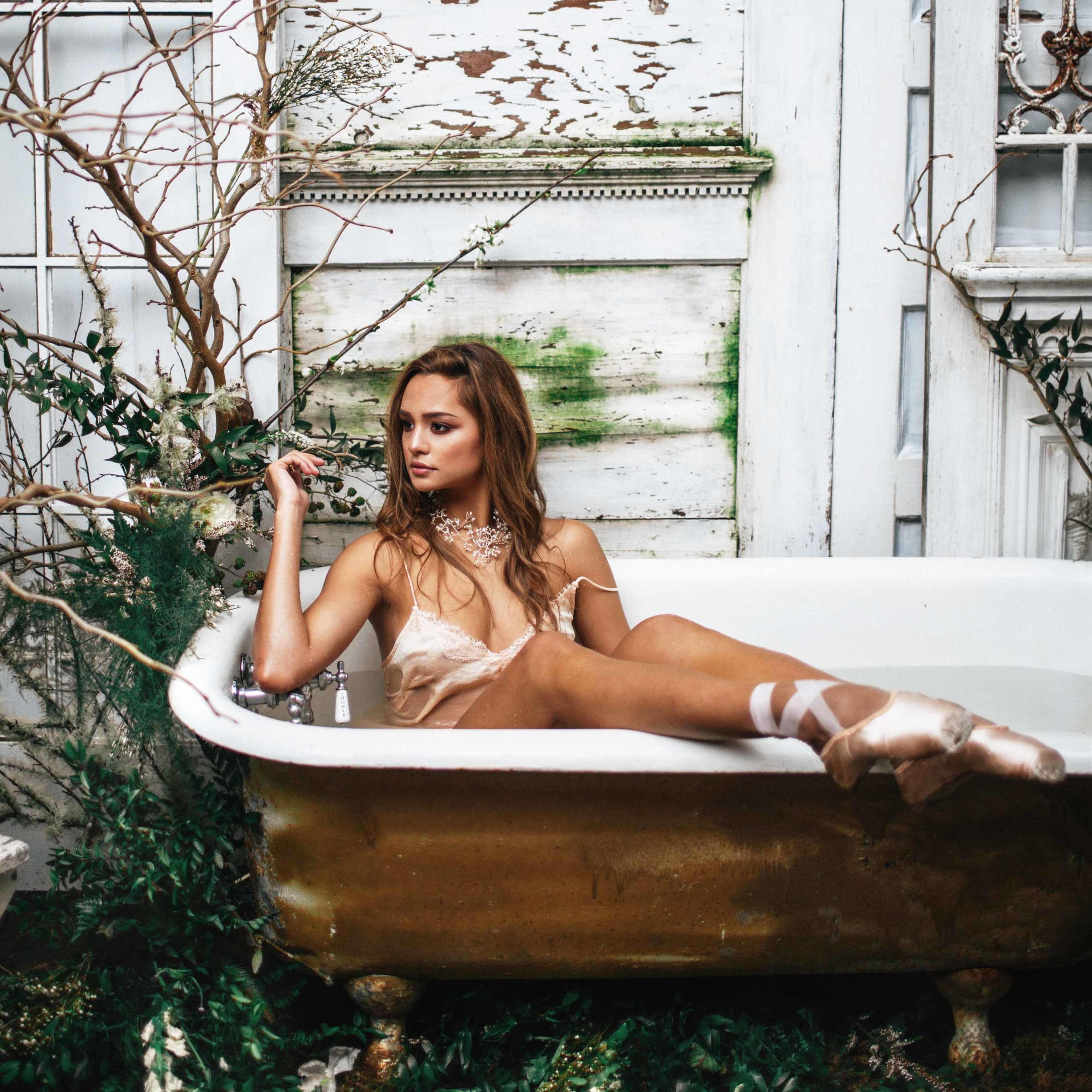 A dancer sits in a vintage bathtub surrounded by greenery and weathered walls.