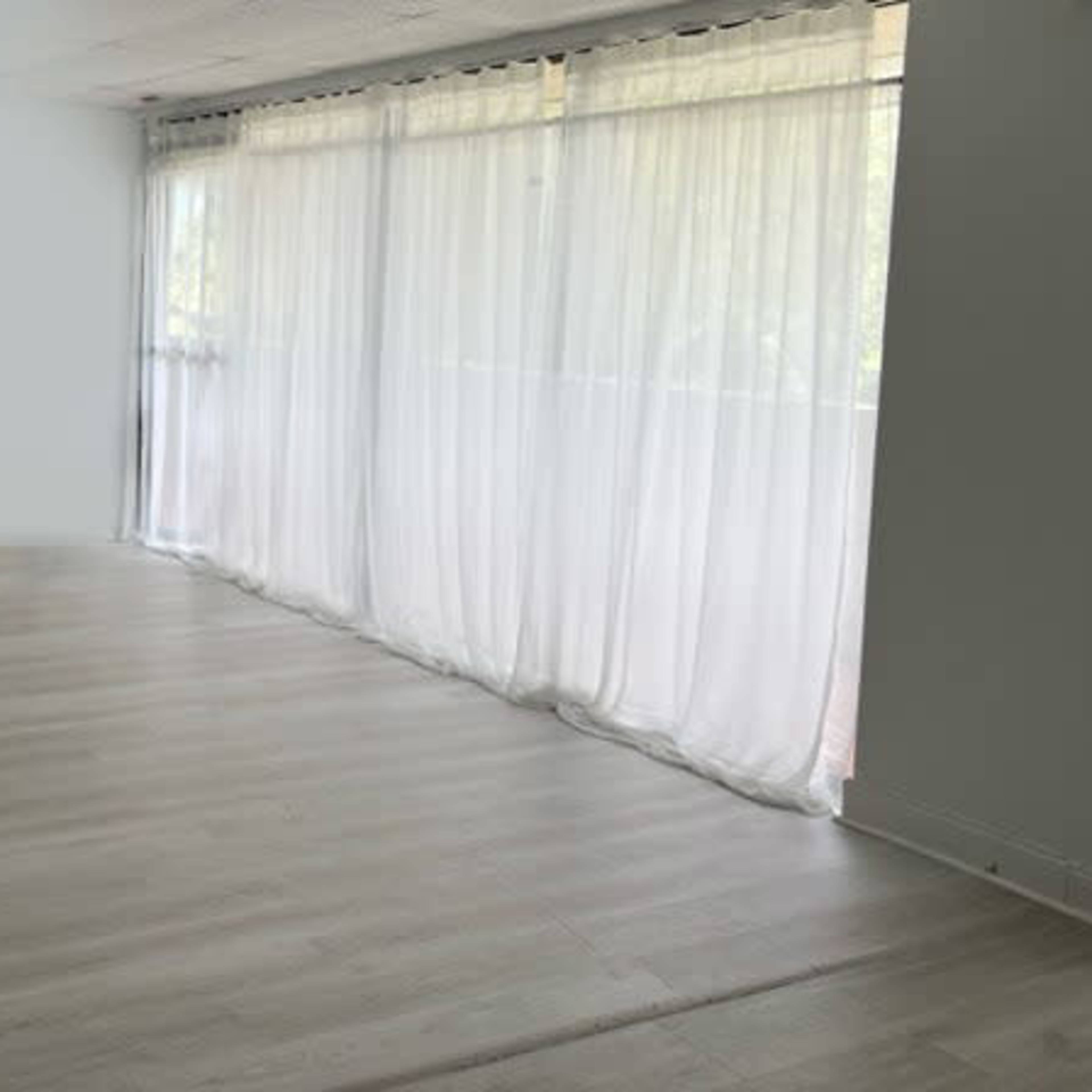 A spacious room with light wooden flooring and sheer curtains covering a large window.