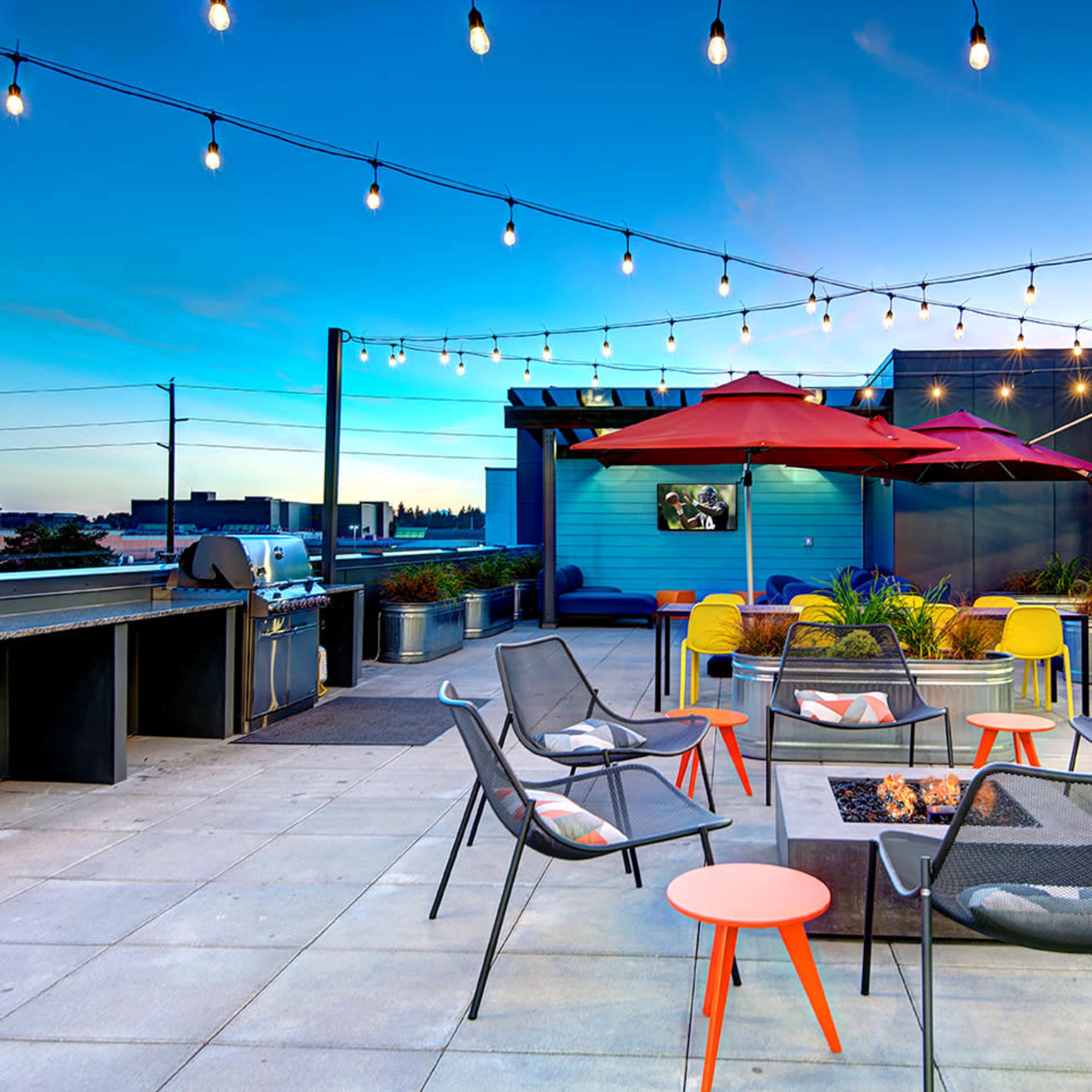 A rooftop terrace adorned with string lights, featuring seating arrangements around a fire pit, tables, and patio umbrellas.