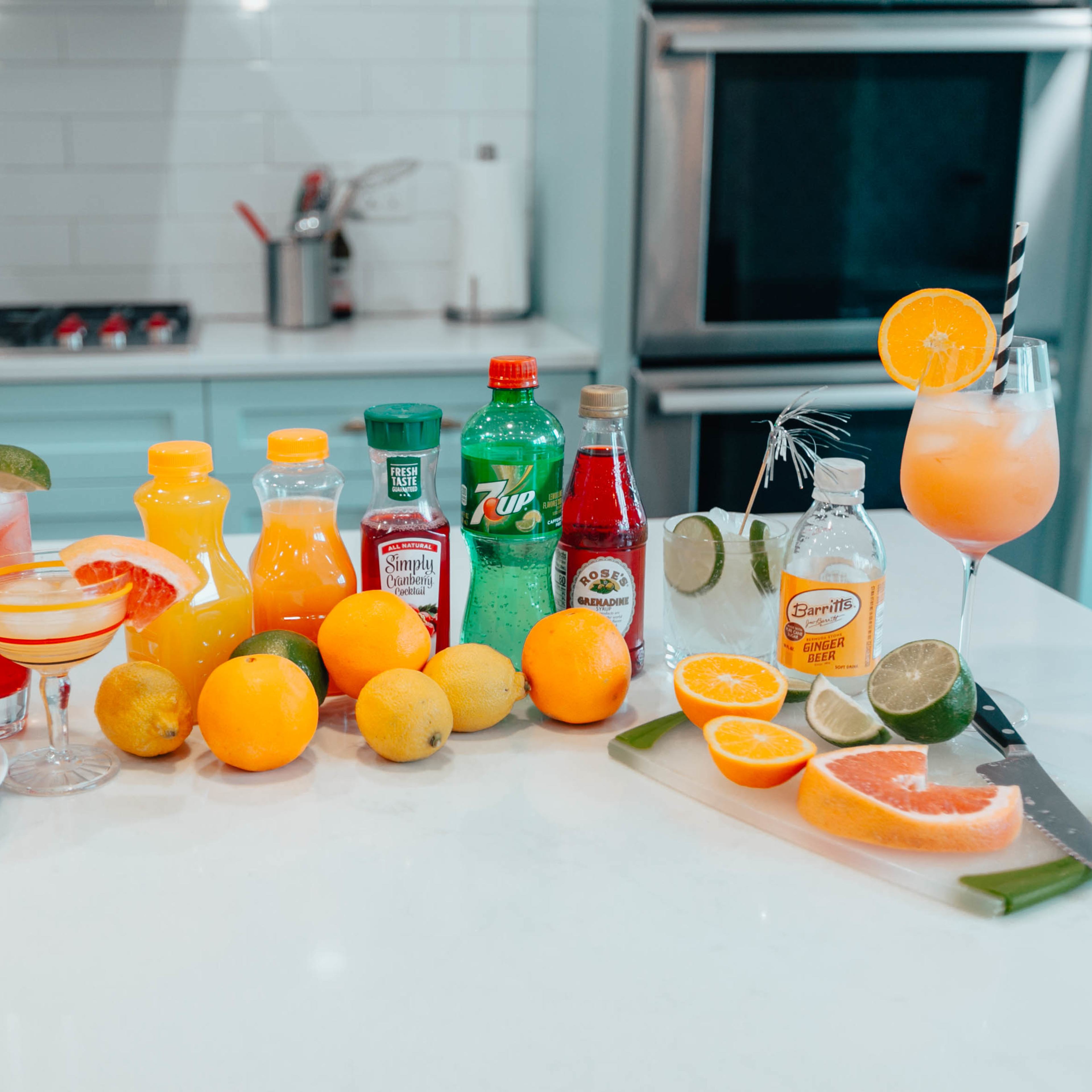 A variety of colorful drinks and fresh fruits arranged on a kitchen countertop, including cocktails, soda bottles, and citrus fruits.