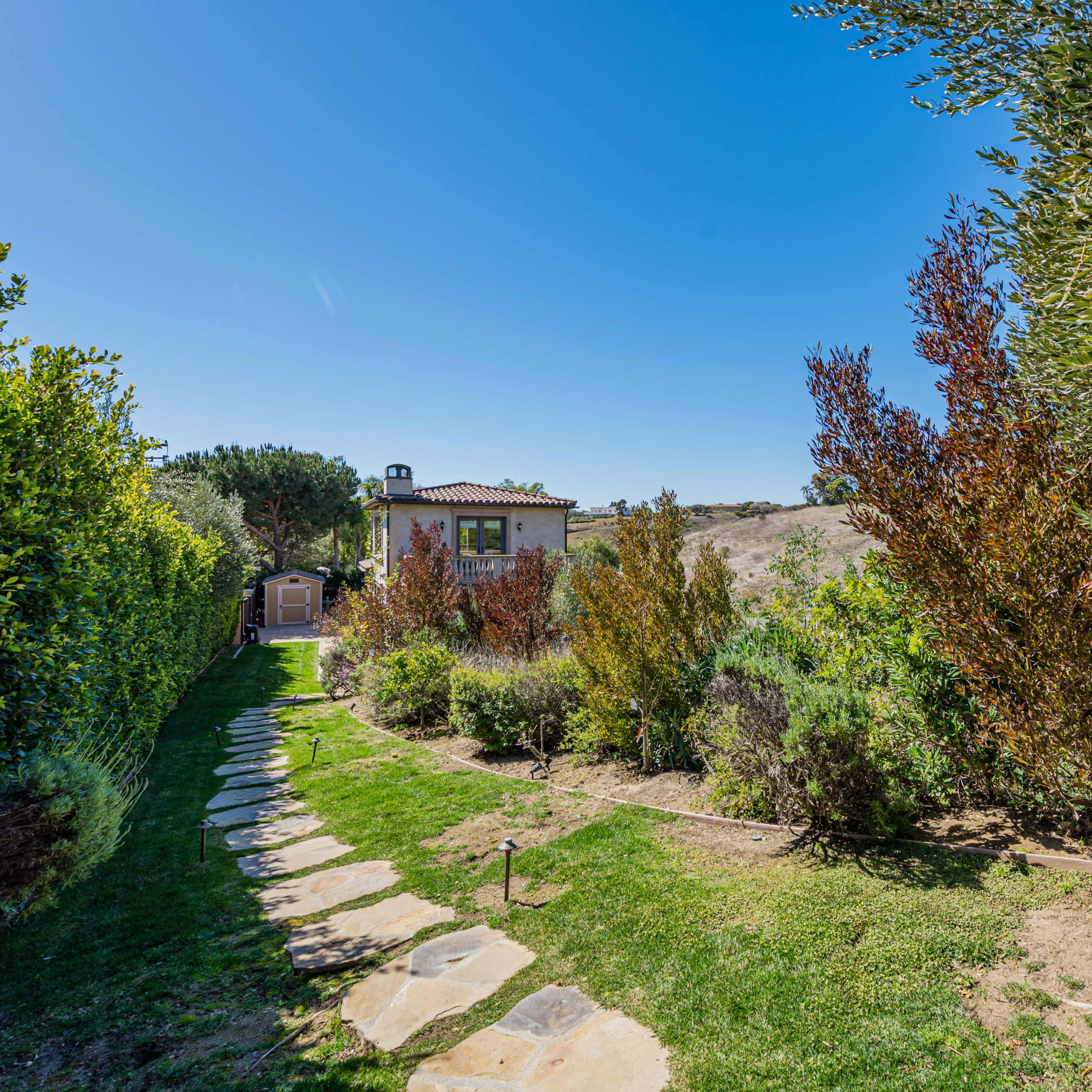 A stone pathway leading through a garden with various shrubs and trees, leading to a small house in the background.