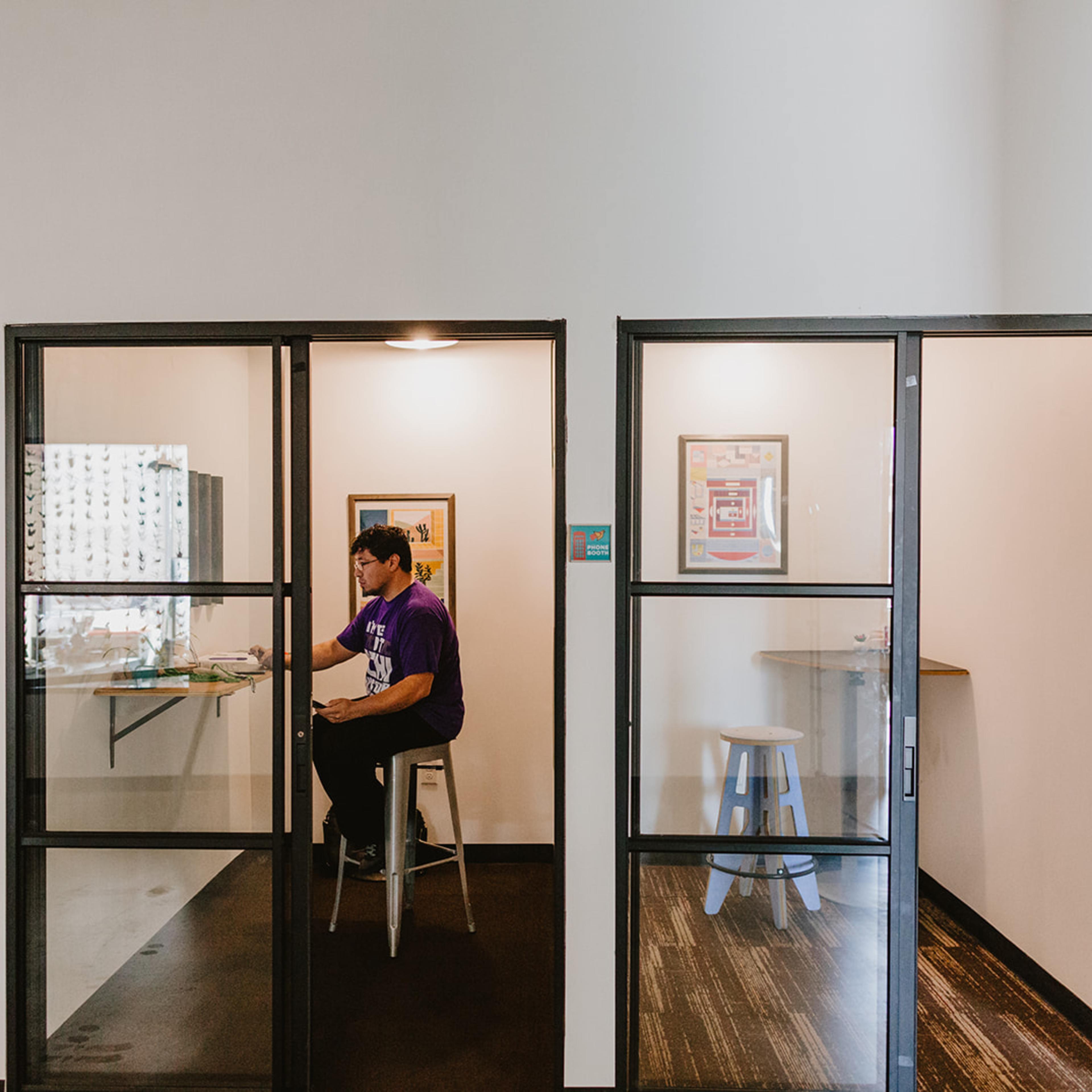 A person sits at a table in an office space, working in a glass-enclosed room beside another empty room with a circular table and stools.