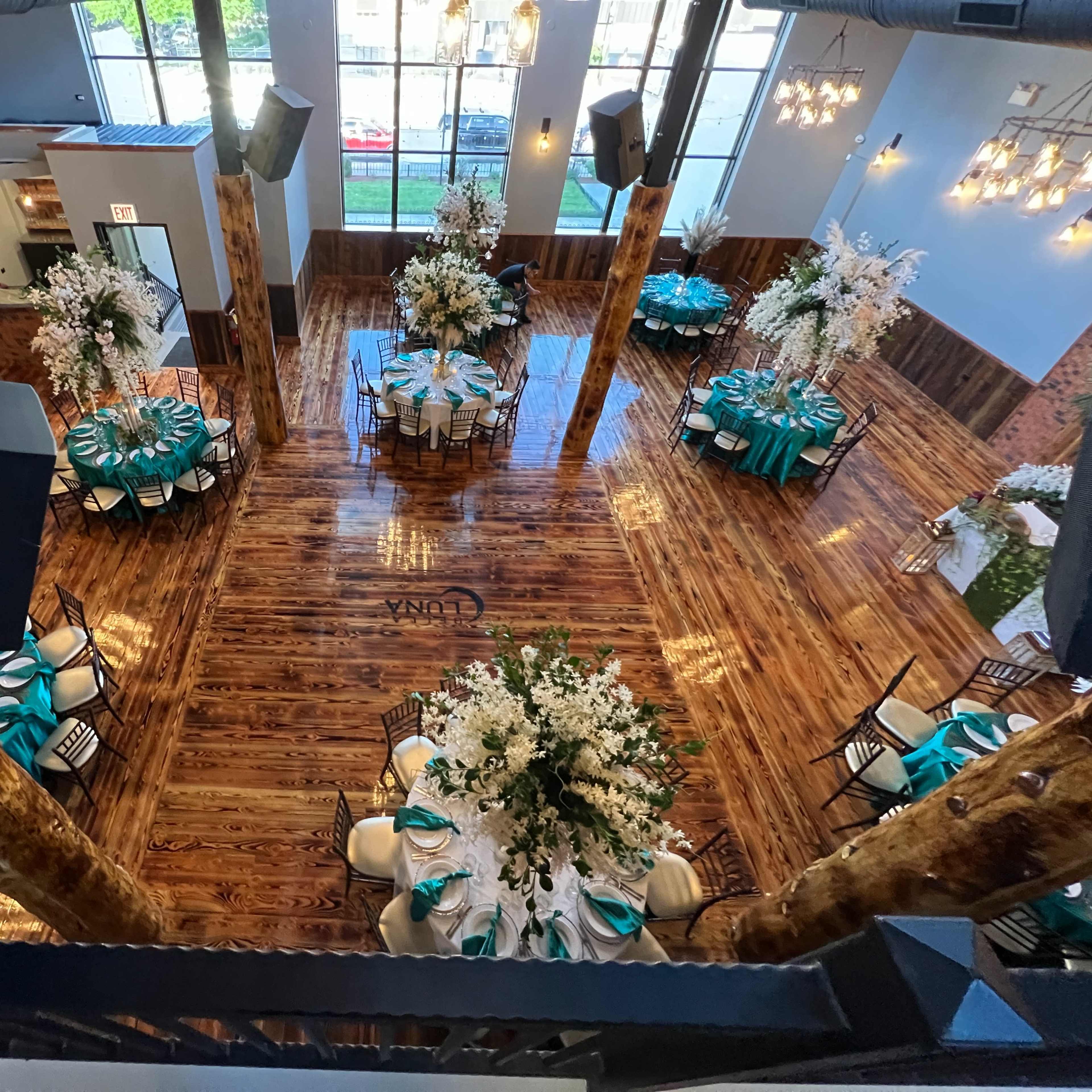 An elegantly arranged event space with tables set and decorated with turquoise and white accents, surrounded by tall floral centerpieces and wooden pillars.