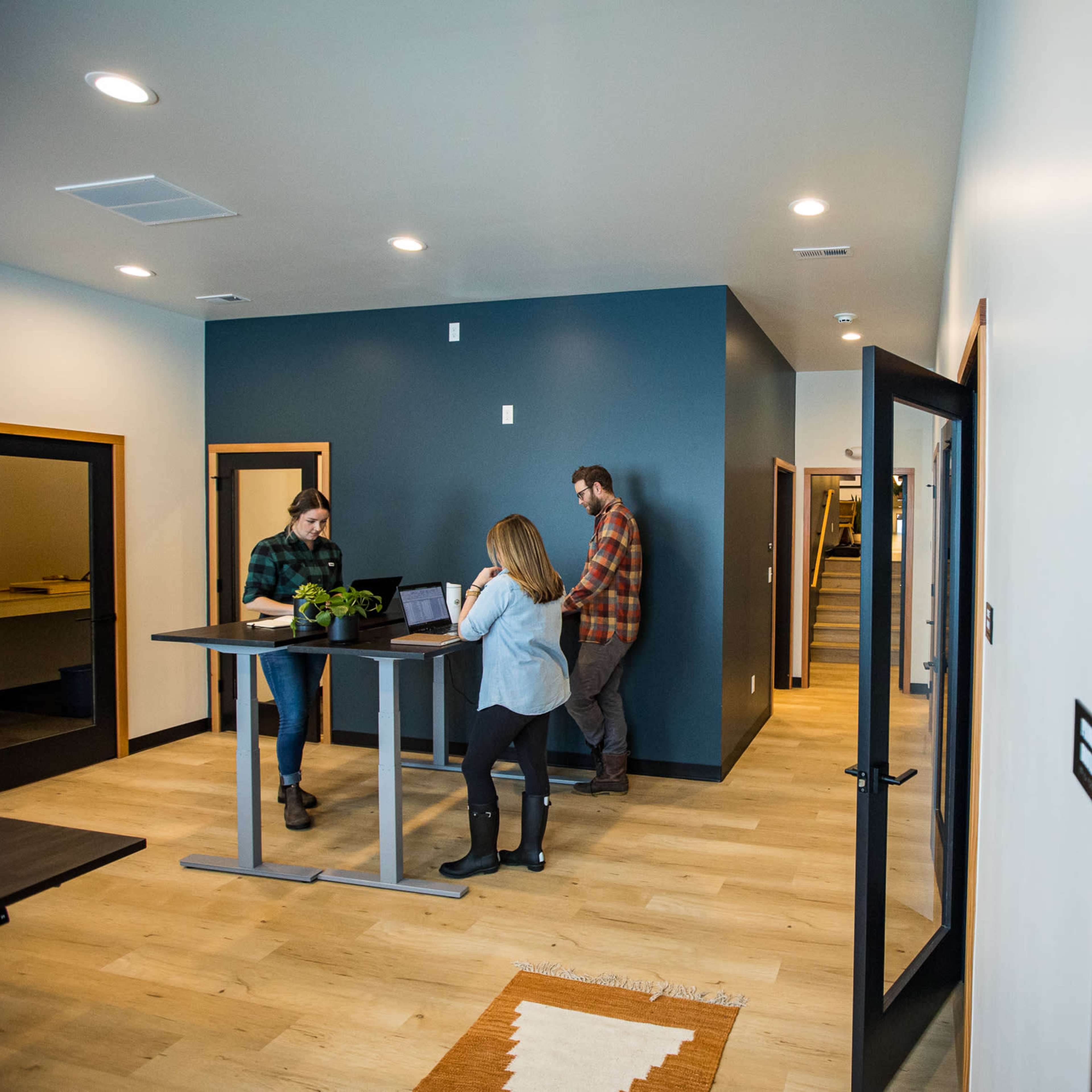 A modern office space with three people engaging in work at standing desks, surrounded by neutral-colored walls and wooden flooring.