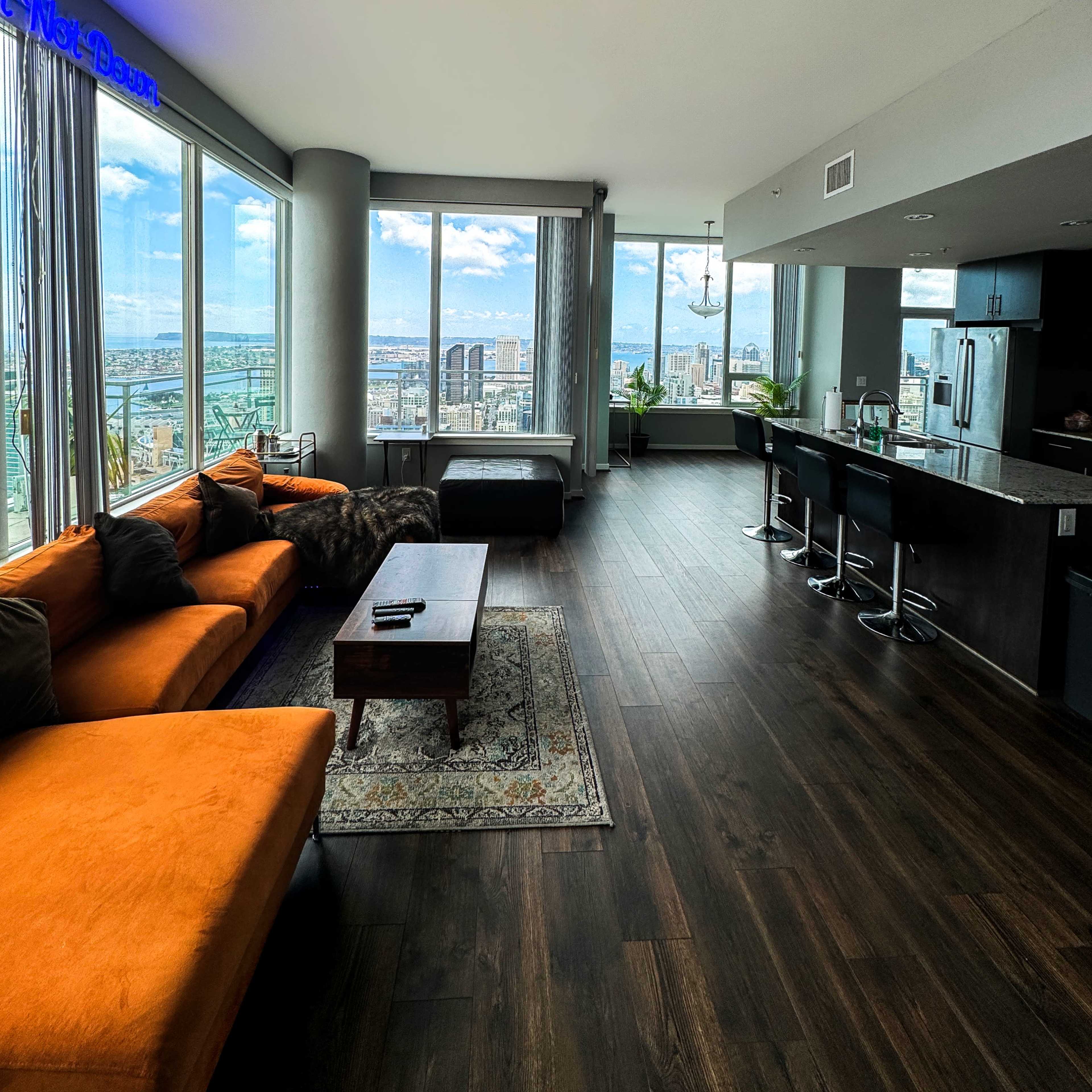 A modern high-rise apartment features an open living area with large windows showcasing a city view, orange couch, and a kitchen bar with stools.