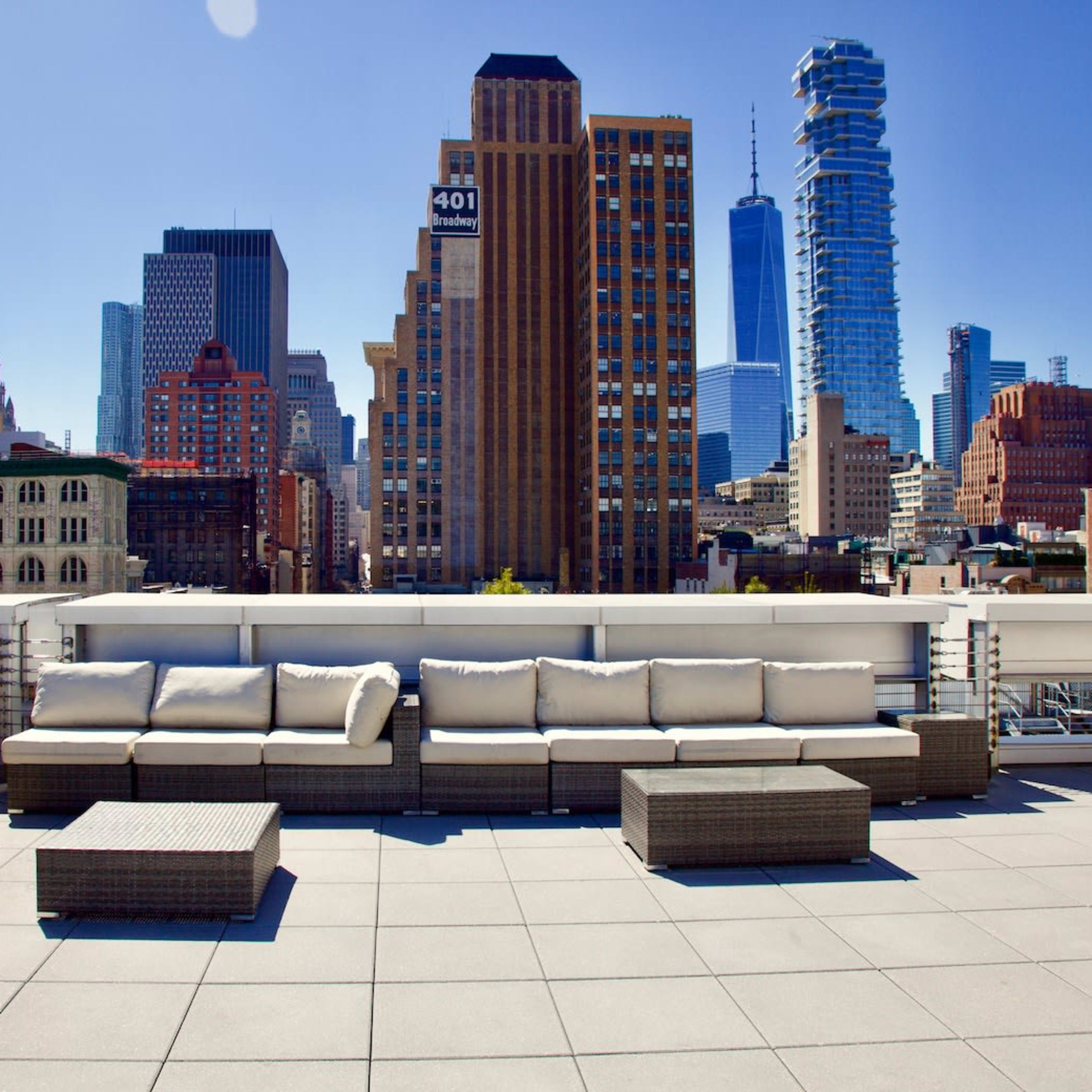 A rooftop terrace features a sectional seating arrangement and a coffee table, overlooking a skyline of tall buildings under a clear blue sky.
