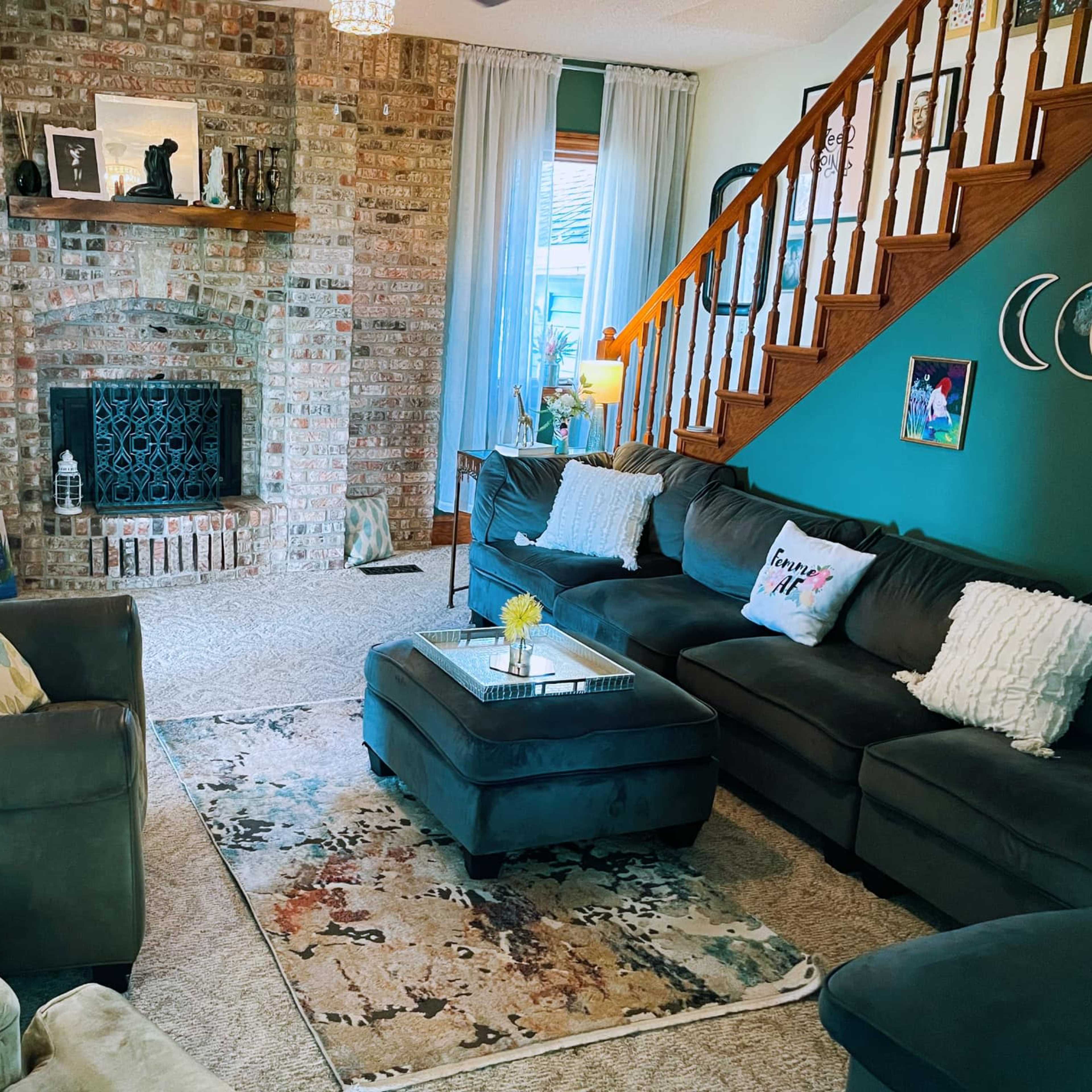 A cozy living room featuring a brick fireplace, a mix of dark-colored sofas, a decorative rug, and a staircase in the background.