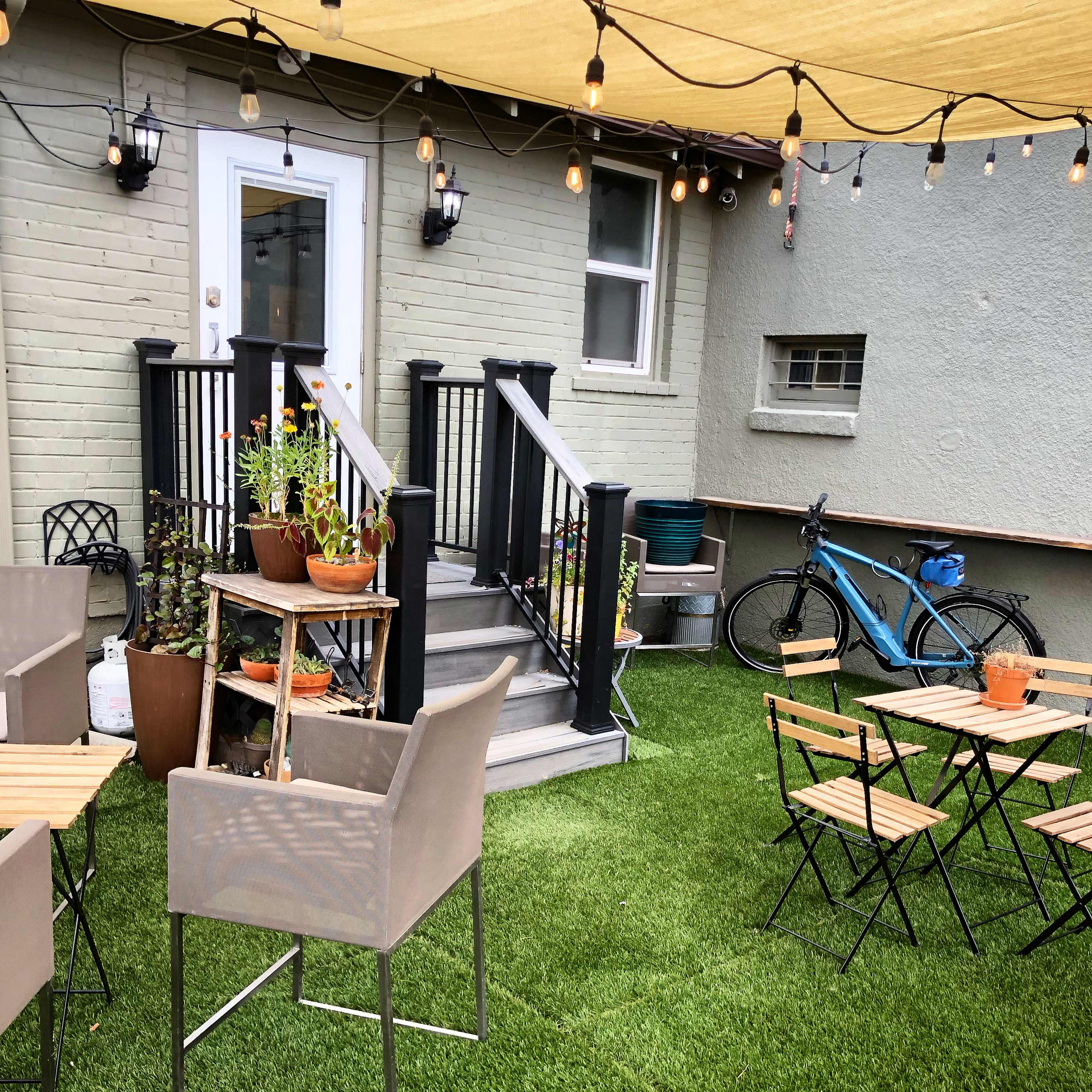 A cozy outdoor patio features green artificial grass, several wooden tables and chairs, potted plants, and a bicycle parked nearby under string lights.