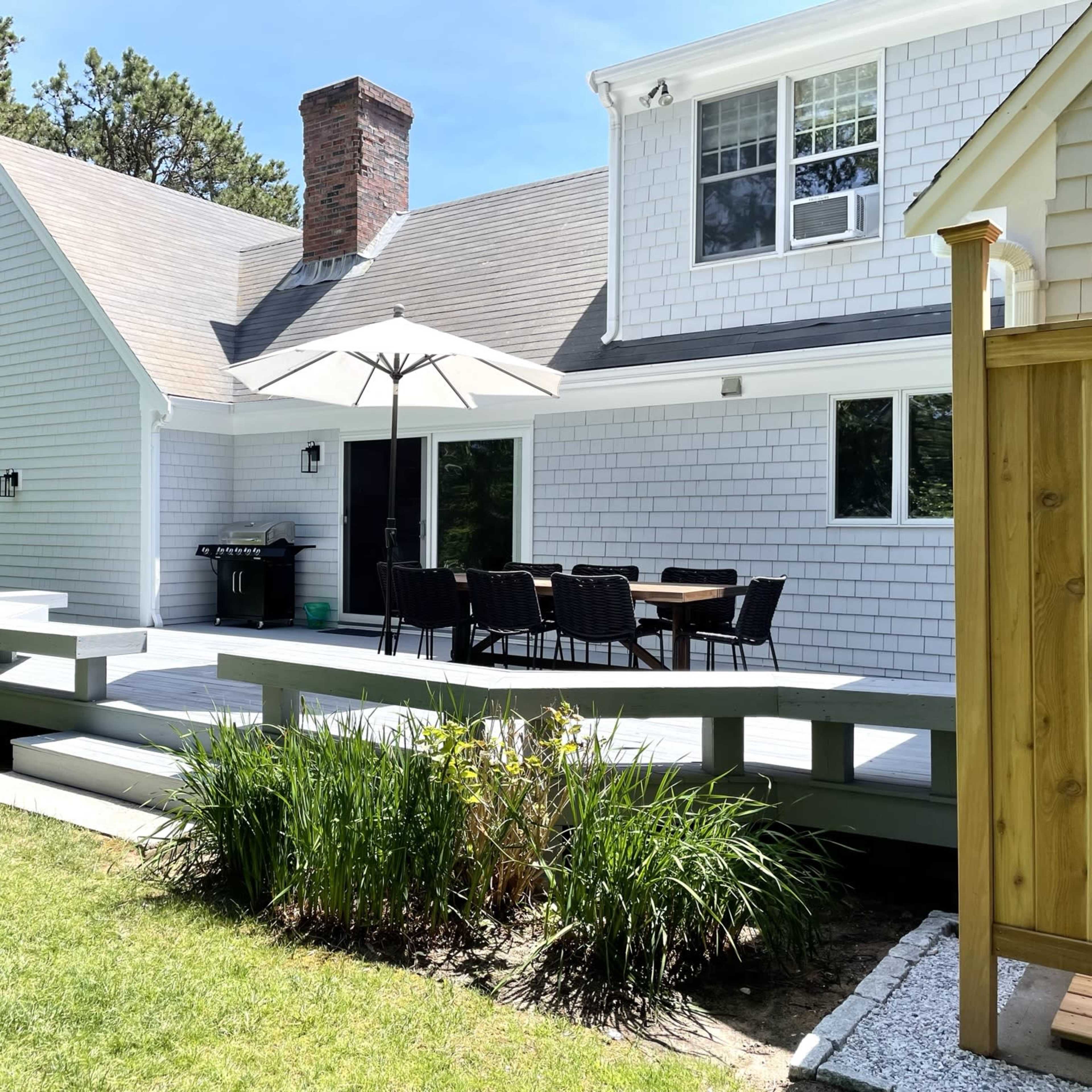 A modern house features a wooden deck with a dining area shaded by an umbrella, surrounded by a grassy lawn and trees.