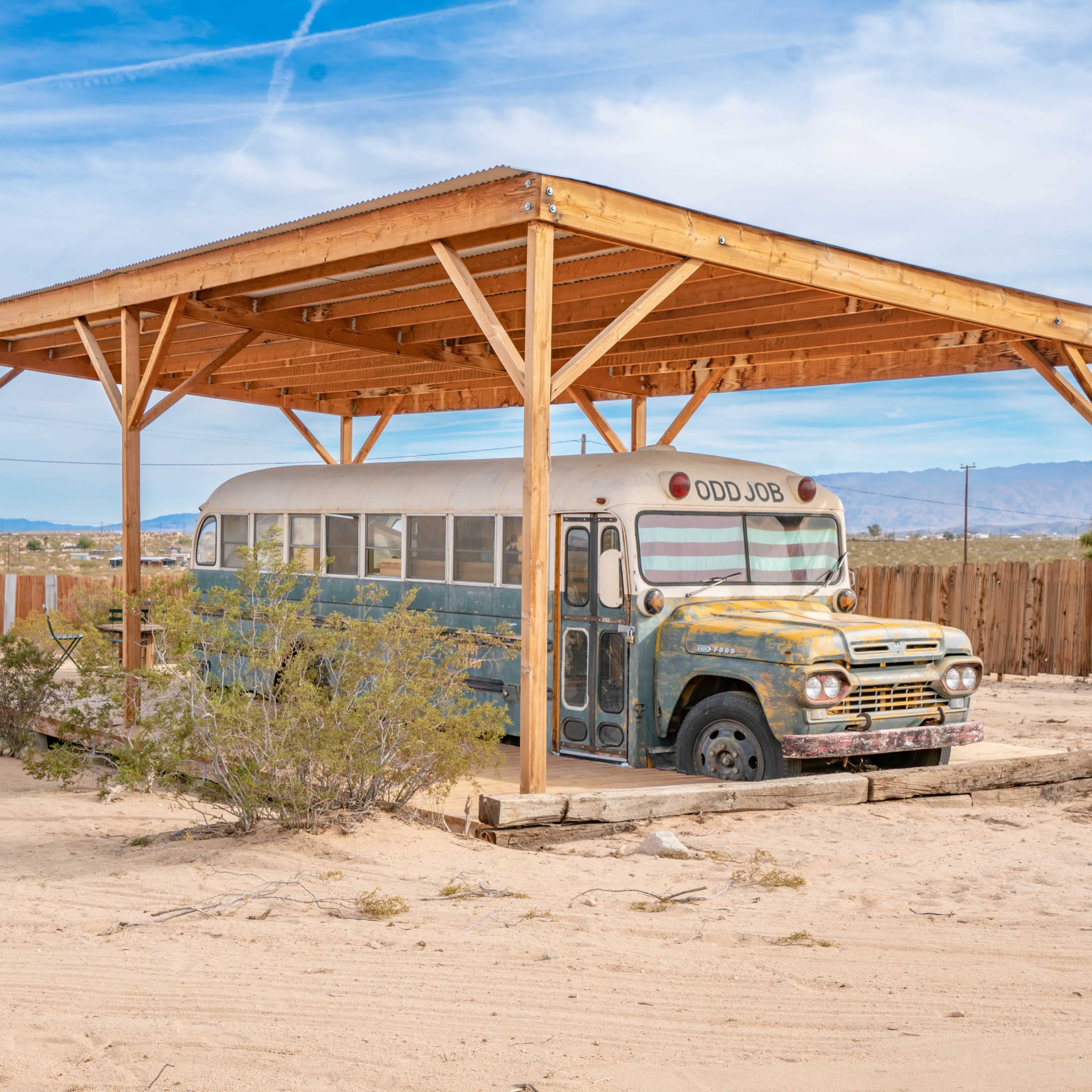 A vintage school bus is parked under a wooden shelter in a desert landscape.