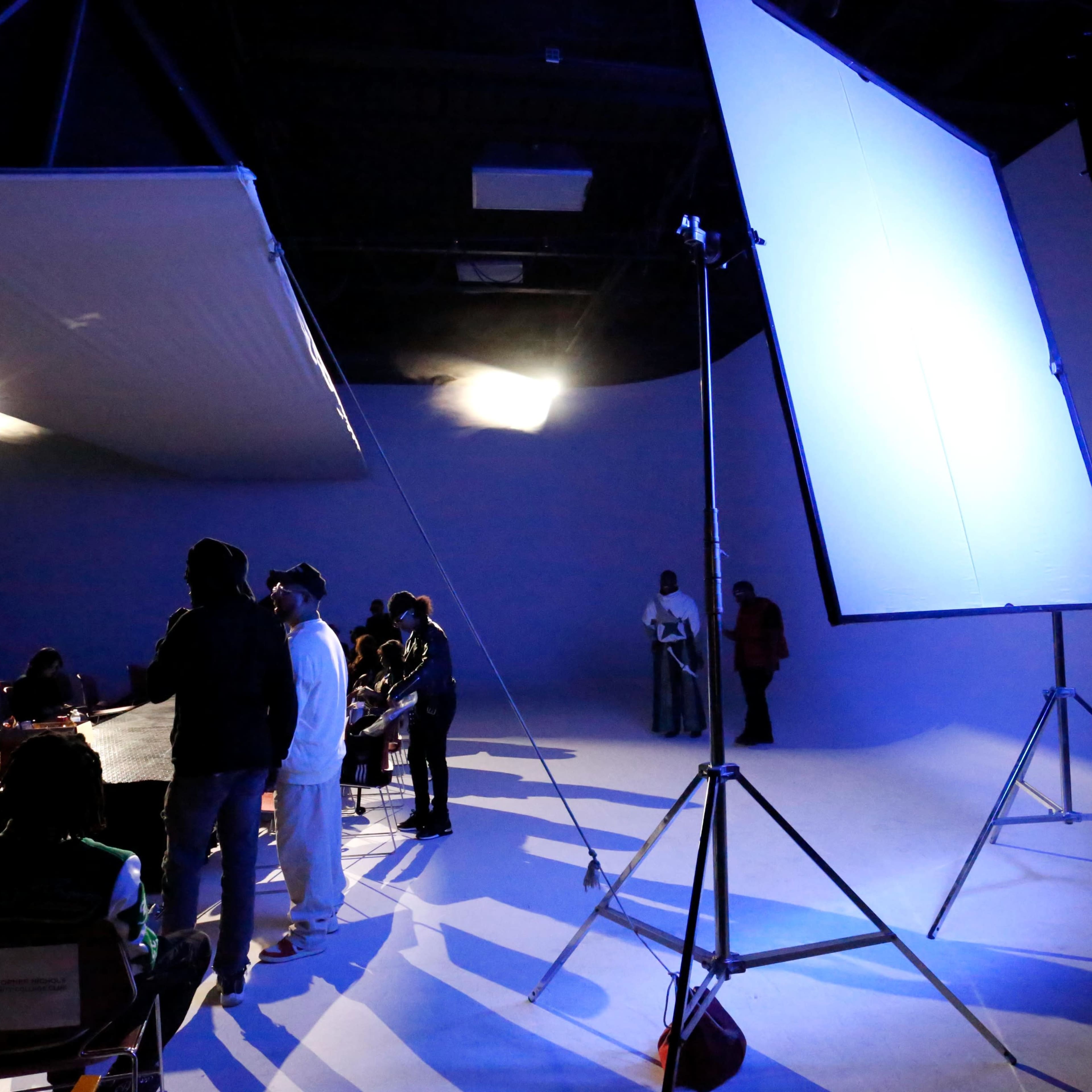A film set is illuminated by large lighting panels, with a group of people gathered around a table in a spacious studio.