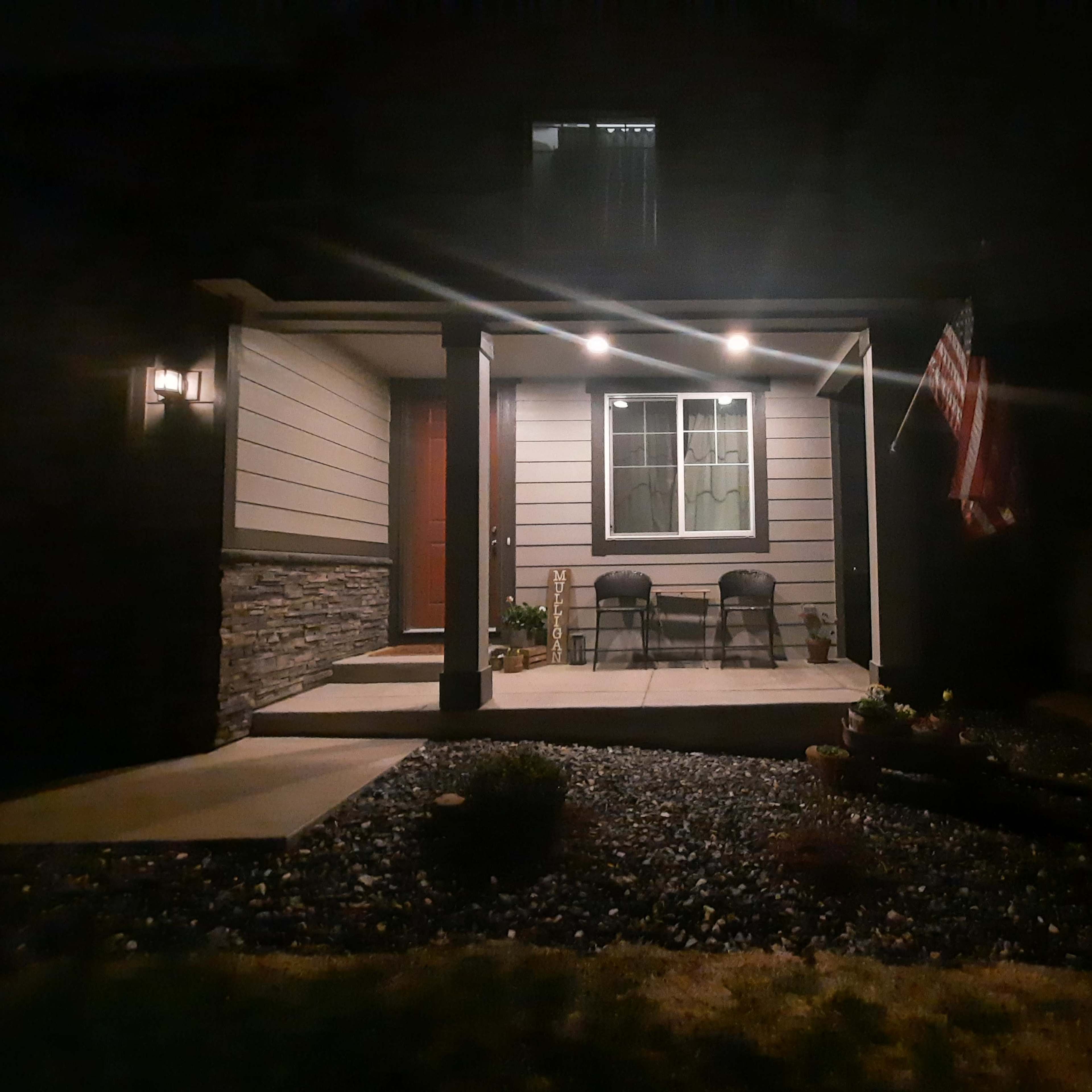 A front porch with two chairs, a door, and an American flag is illuminated at night.