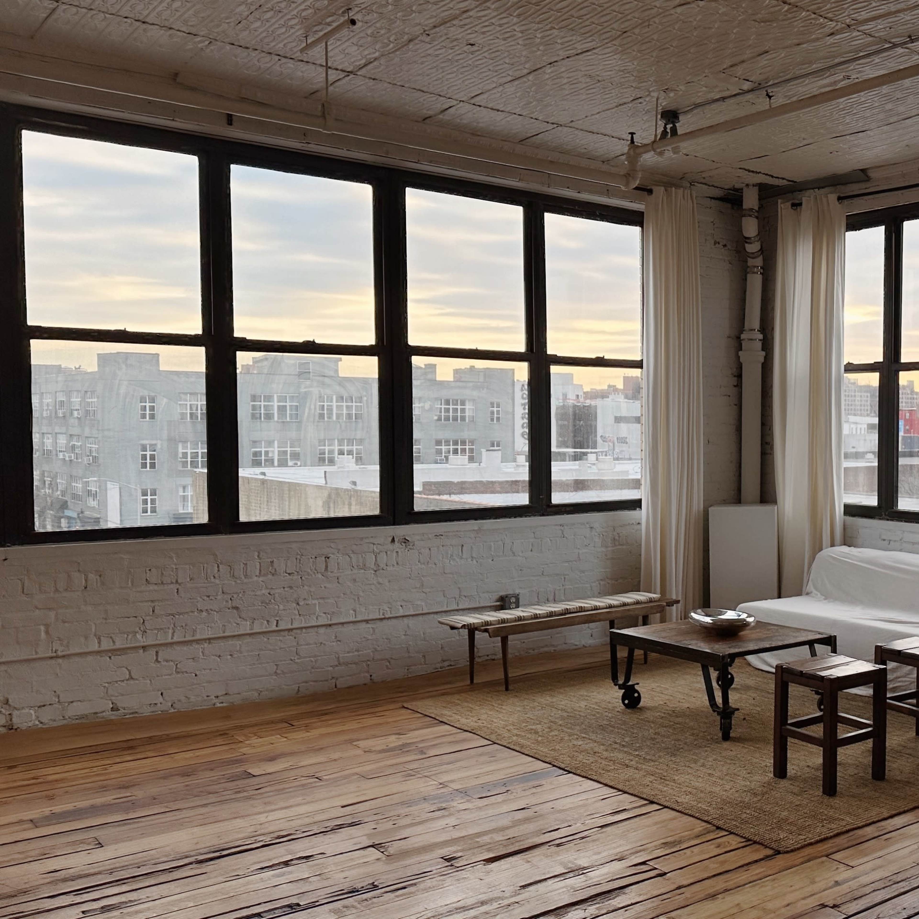 A spacious, minimalist room with large windows, a white sofa, a rug, a wooden coffee table, and a tall mirror.