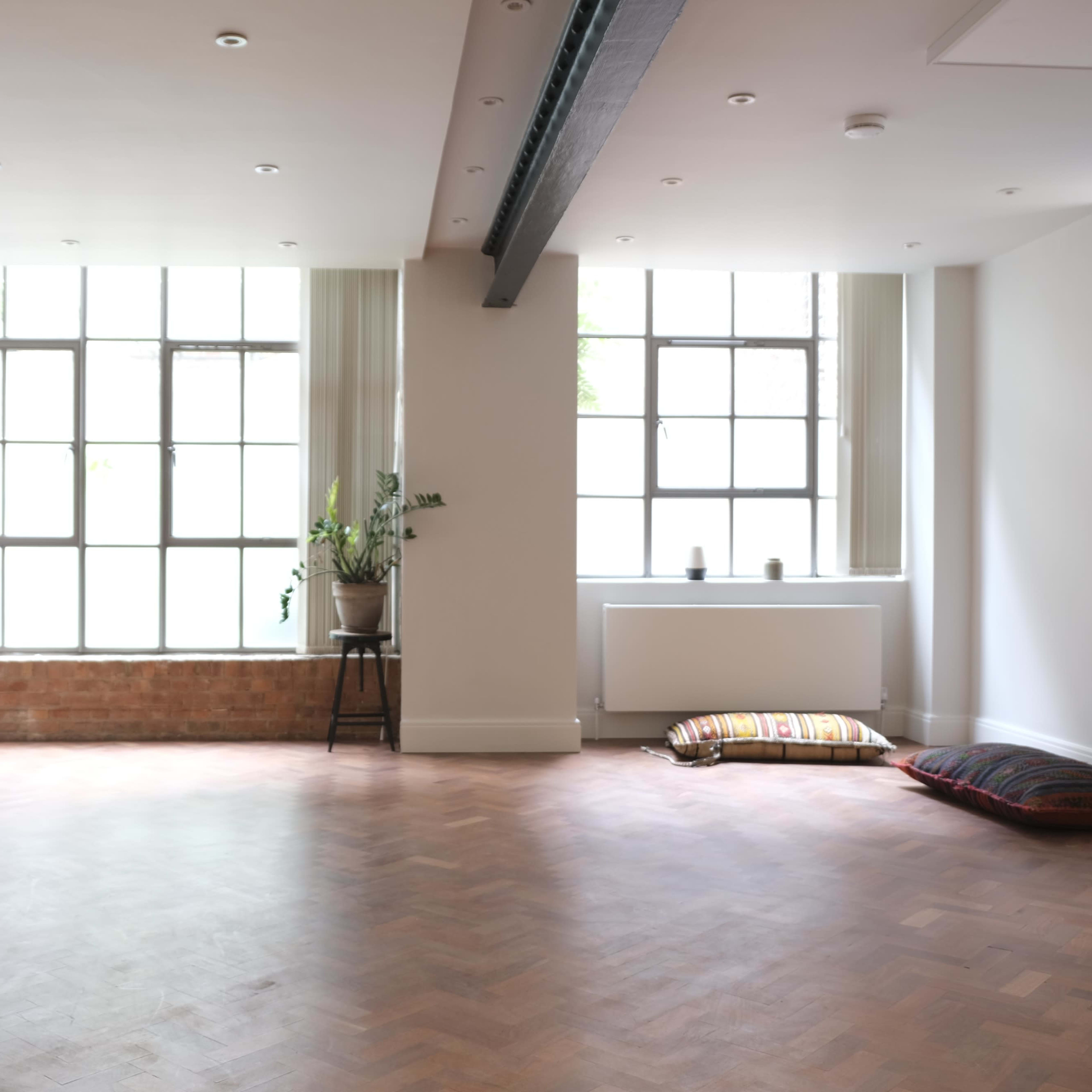 A spacious, well-lit room with large windows, hardwood flooring, and minimal furnishings, including a potted plant and floor cushions.
