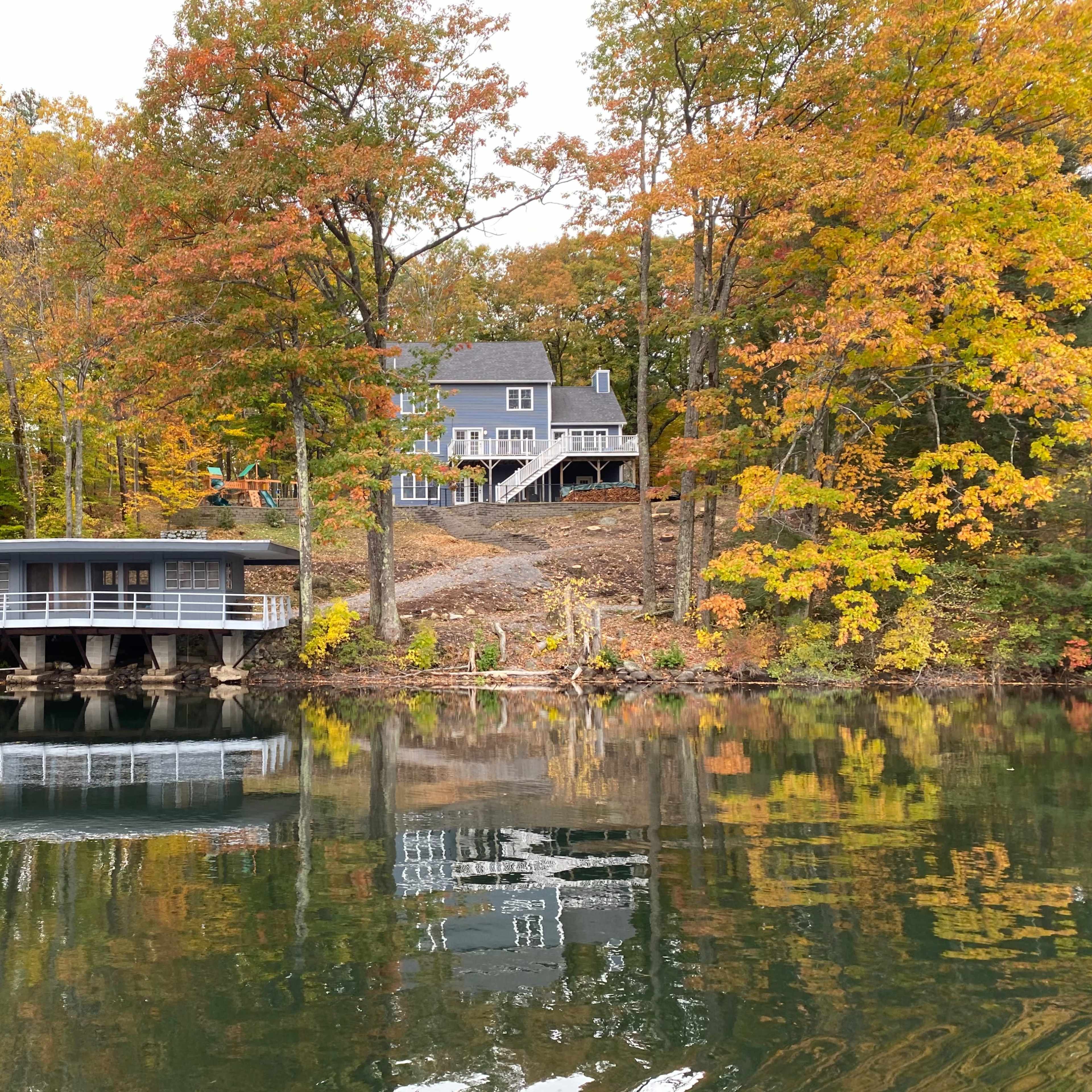 A serene lakeside scene features two houses with autumn foliage reflecting on the water's surface.