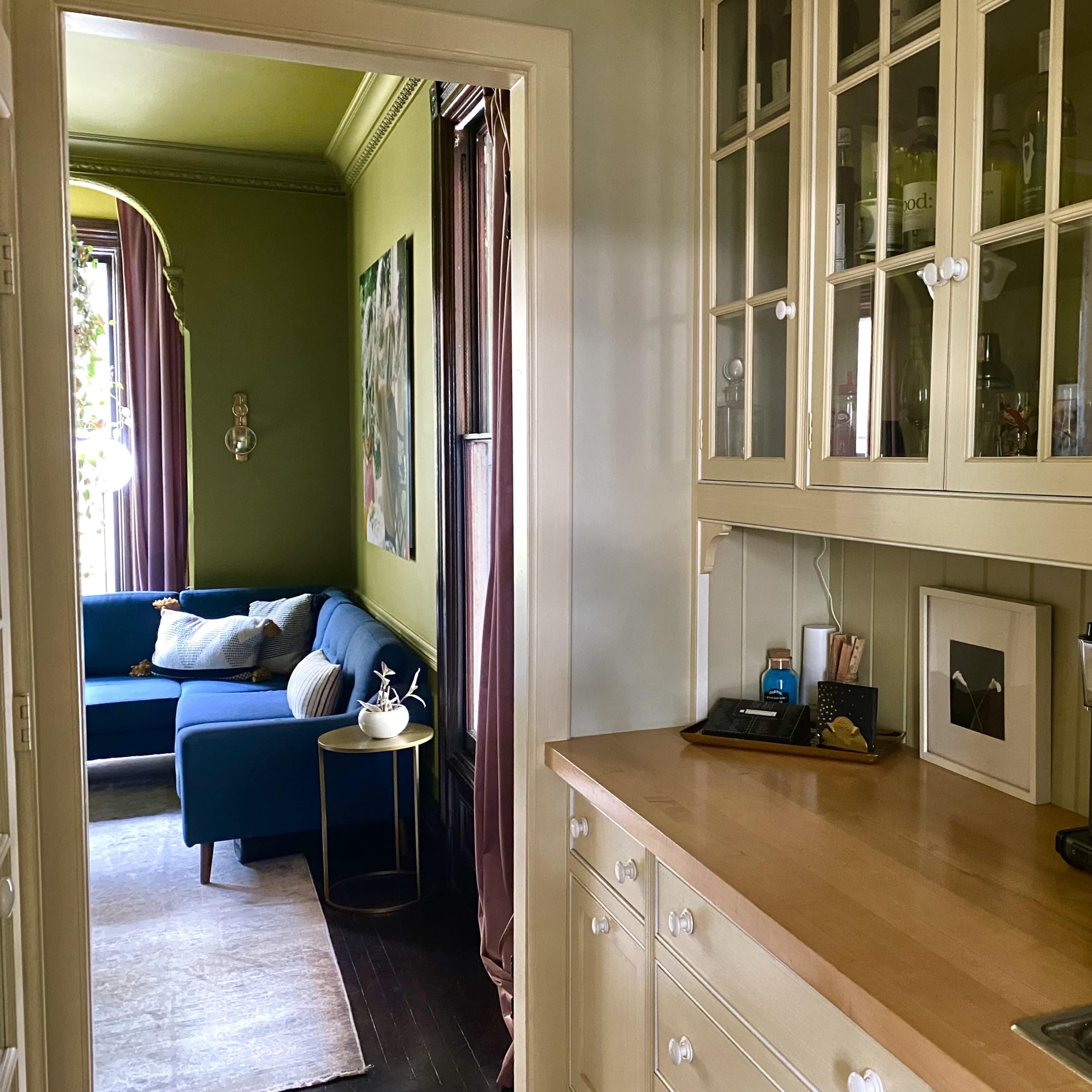 A hallway leading to a living room with a blue couch, framed art on the walls, and a countertop with kitchen appliances and decor.