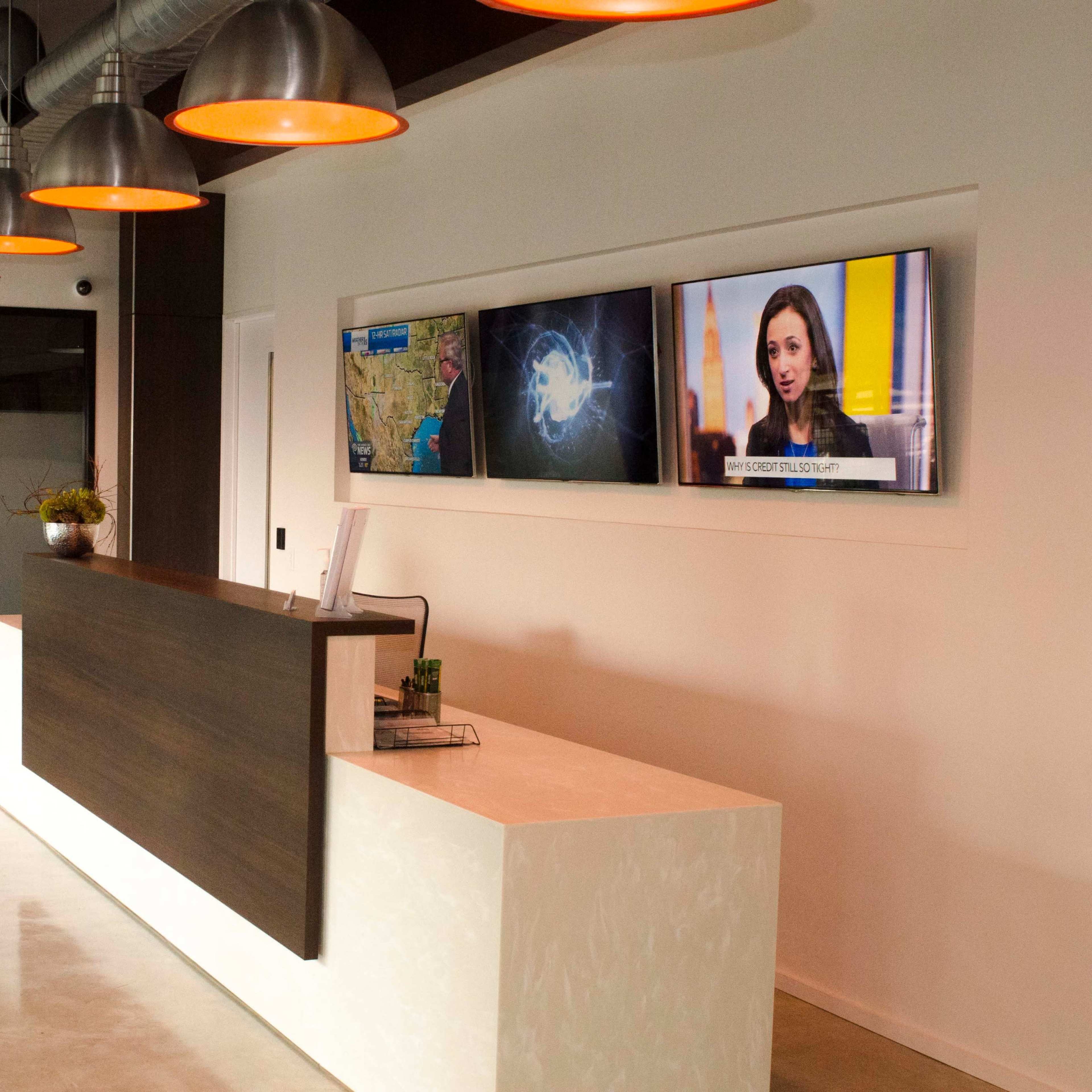 A modern reception area features a white front desk, two large screens displaying information and news, and overhead pendant lighting.