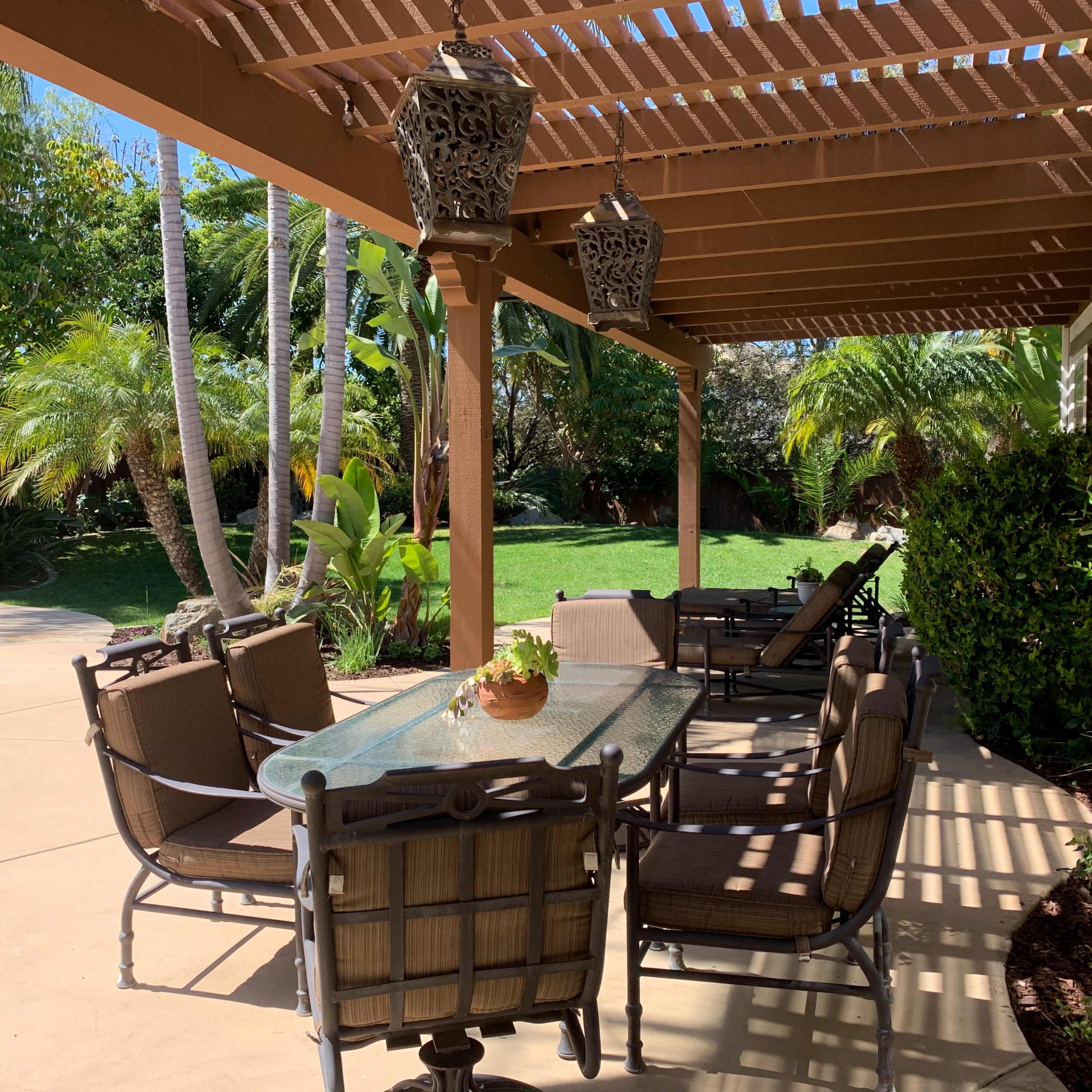 A covered outdoor dining area features a glass table surrounded by chairs, with palm trees and a grassy lawn in the background.