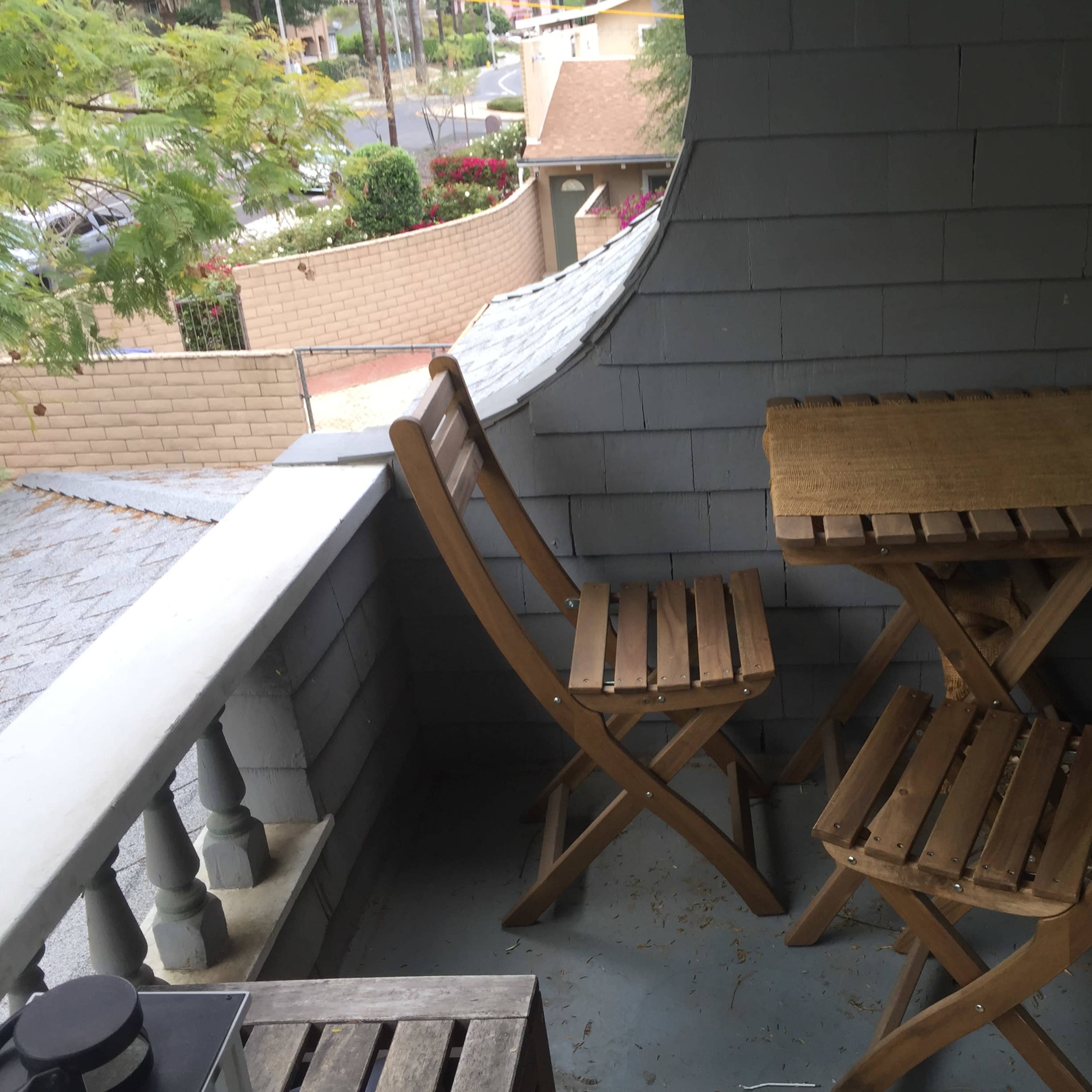 A small balcony with two wooden folding chairs and a matching table, surrounded by a railing and shaded by trees.