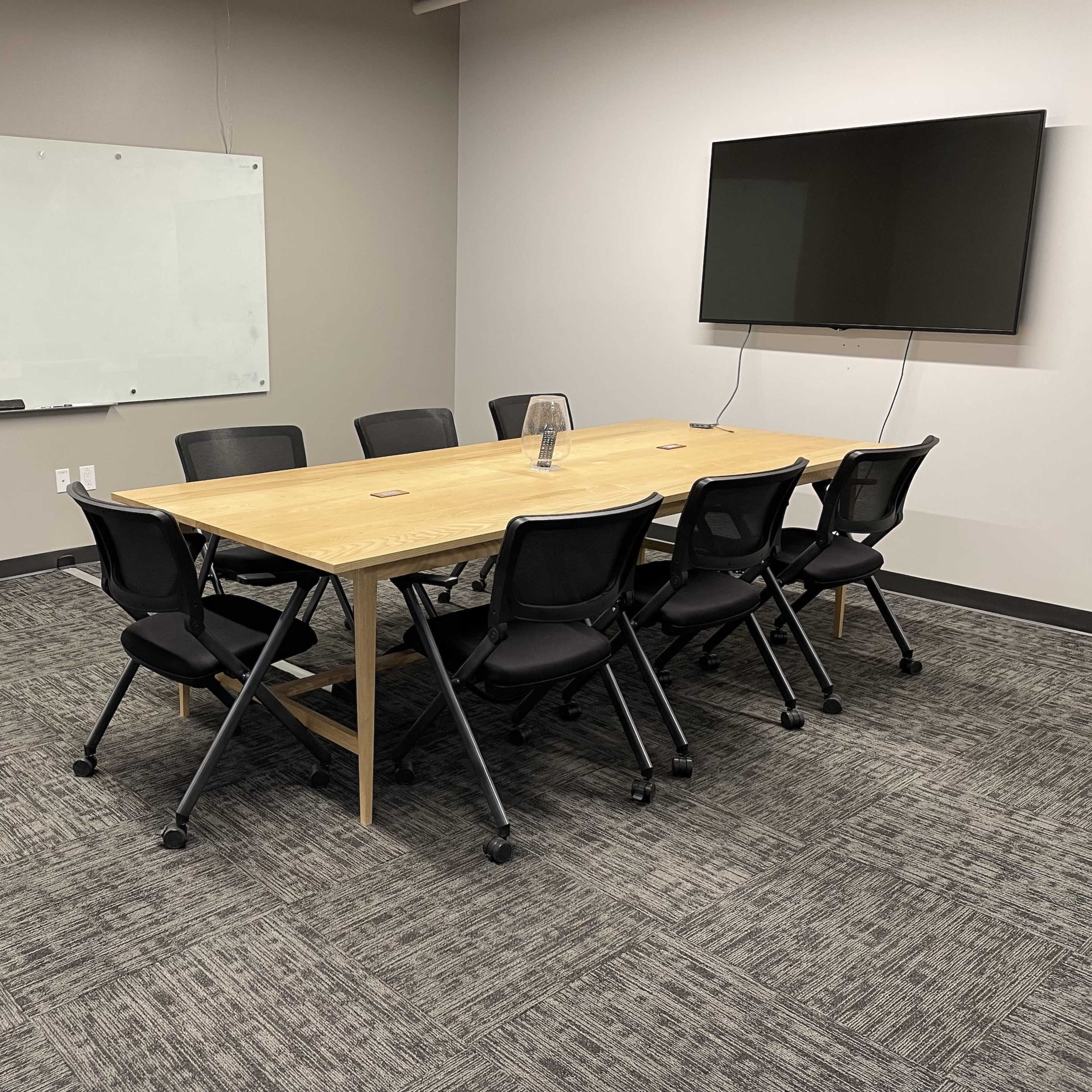 A conference room featuring a long wooden table surrounded by eight black chairs, a whiteboard on the wall, and a mounted television.