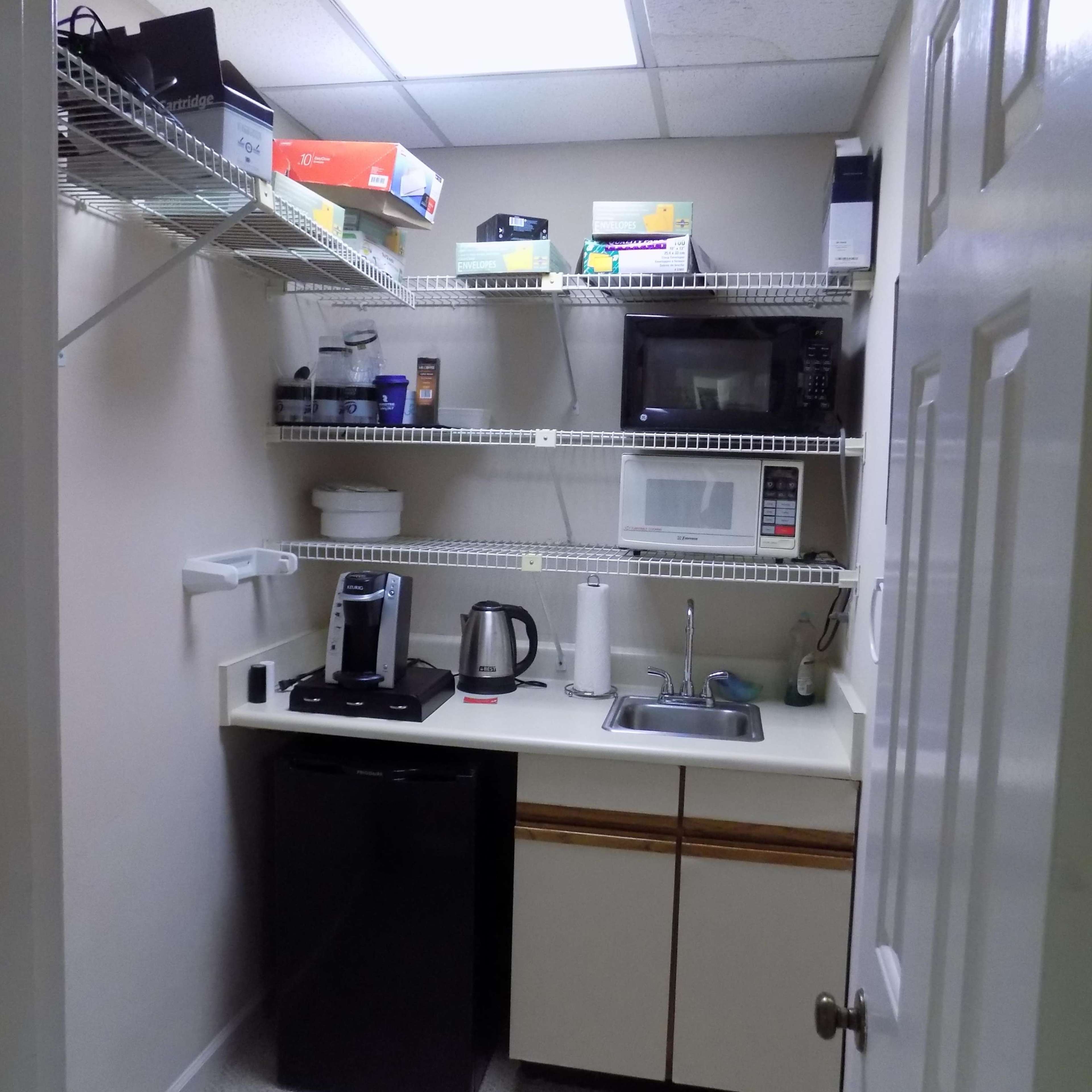 A small kitchen area featuring a sink, a coffee maker, a microwave, and shelves stocked with various appliances and items.