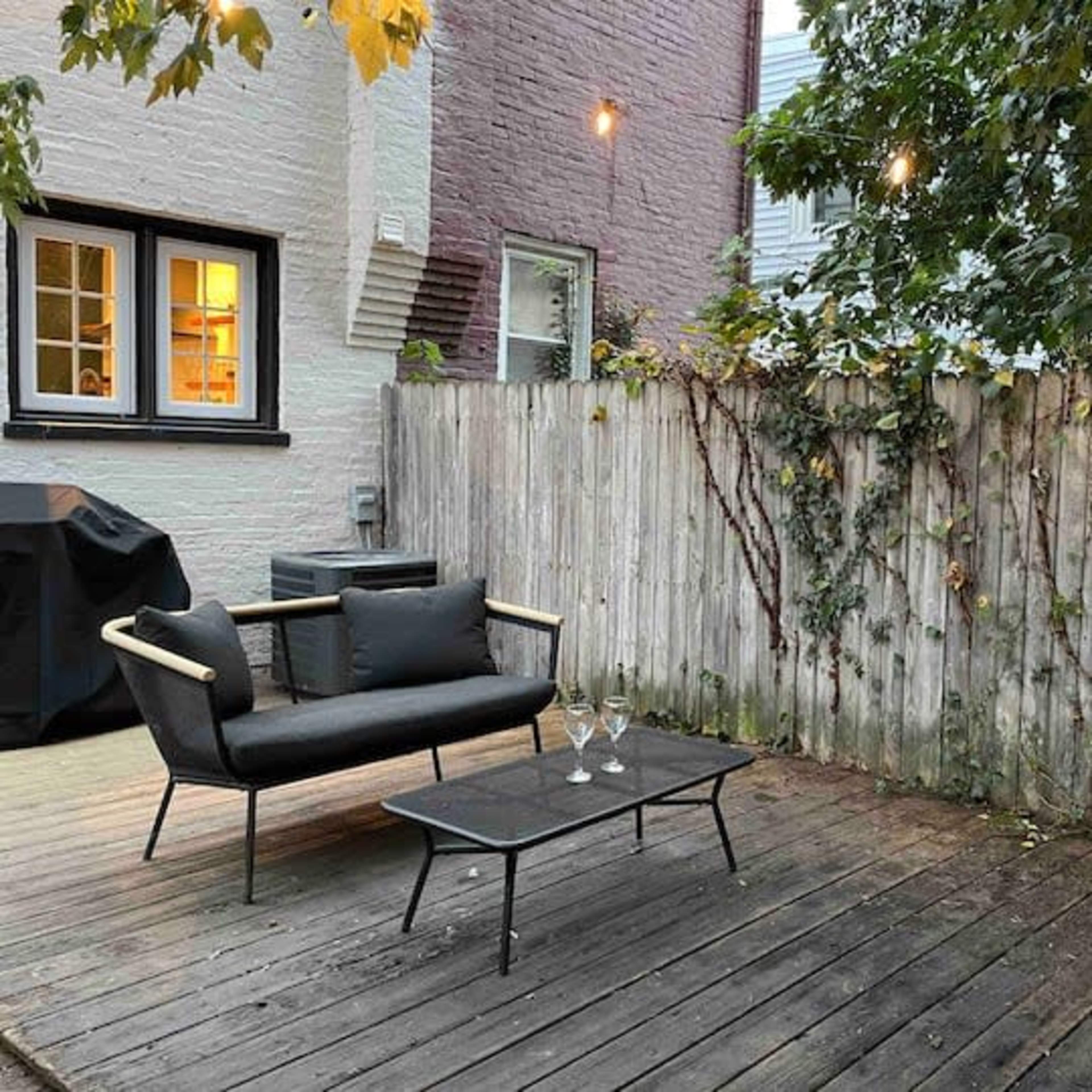 A small outdoor seating area features a black sofa and a glass table on a wooden deck, surrounded by a wooden fence with climbing vines.