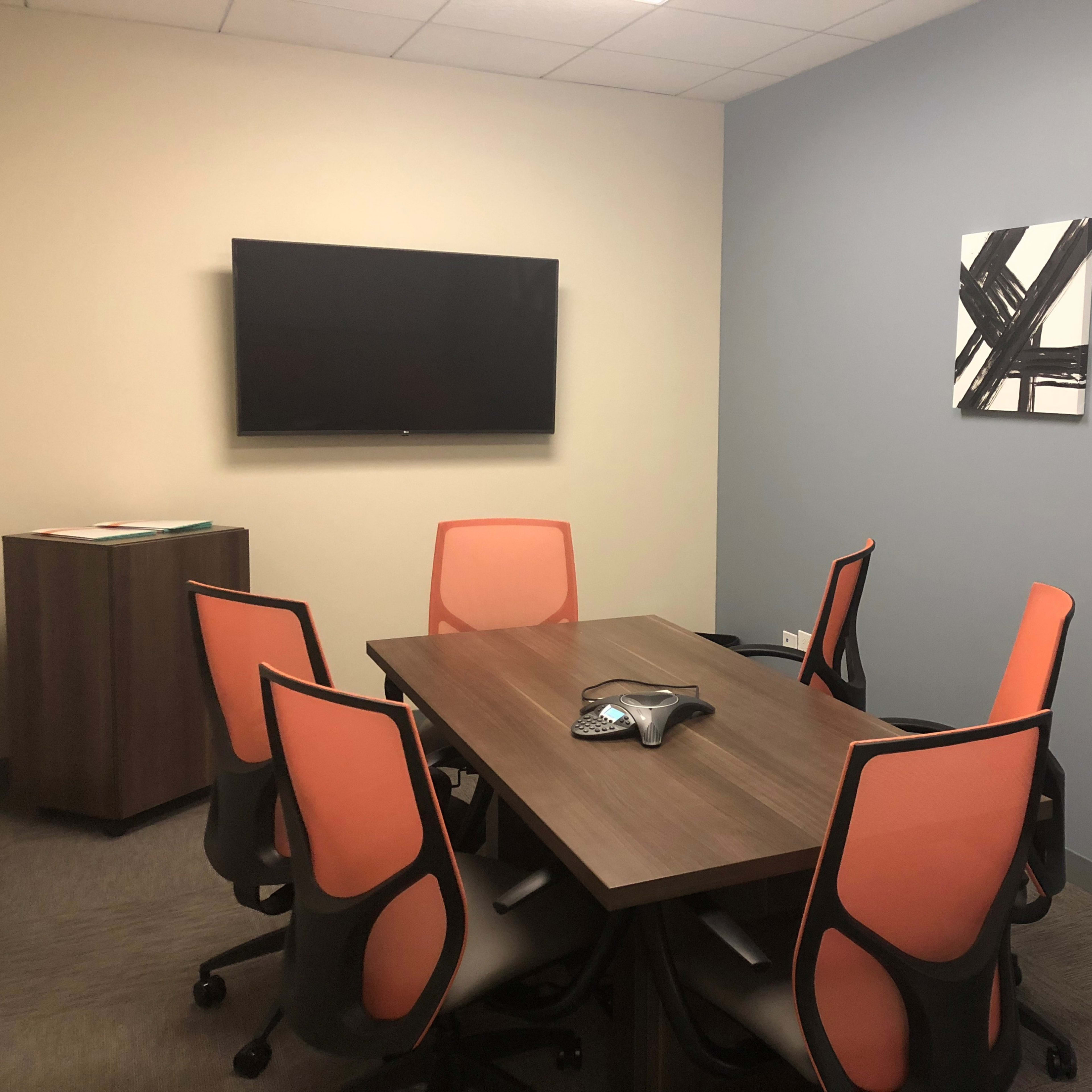 A conference room equipped with a wooden table surrounded by six orange and black chairs, a wall-mounted television, and two abstract framed artworks.