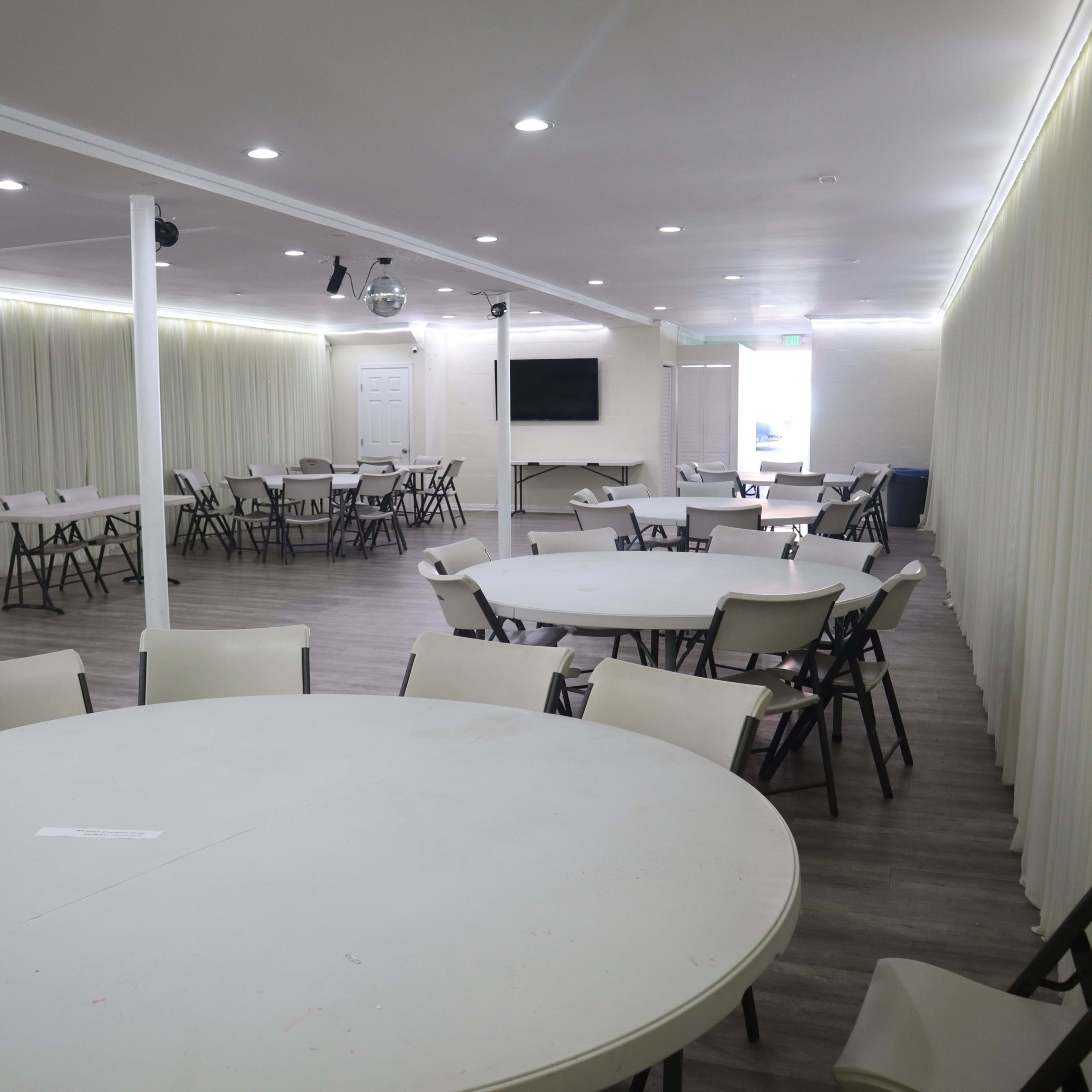An empty event room with round tables and chairs arranged throughout, illuminated by overhead lights and characterized by light-colored curtains along the walls.