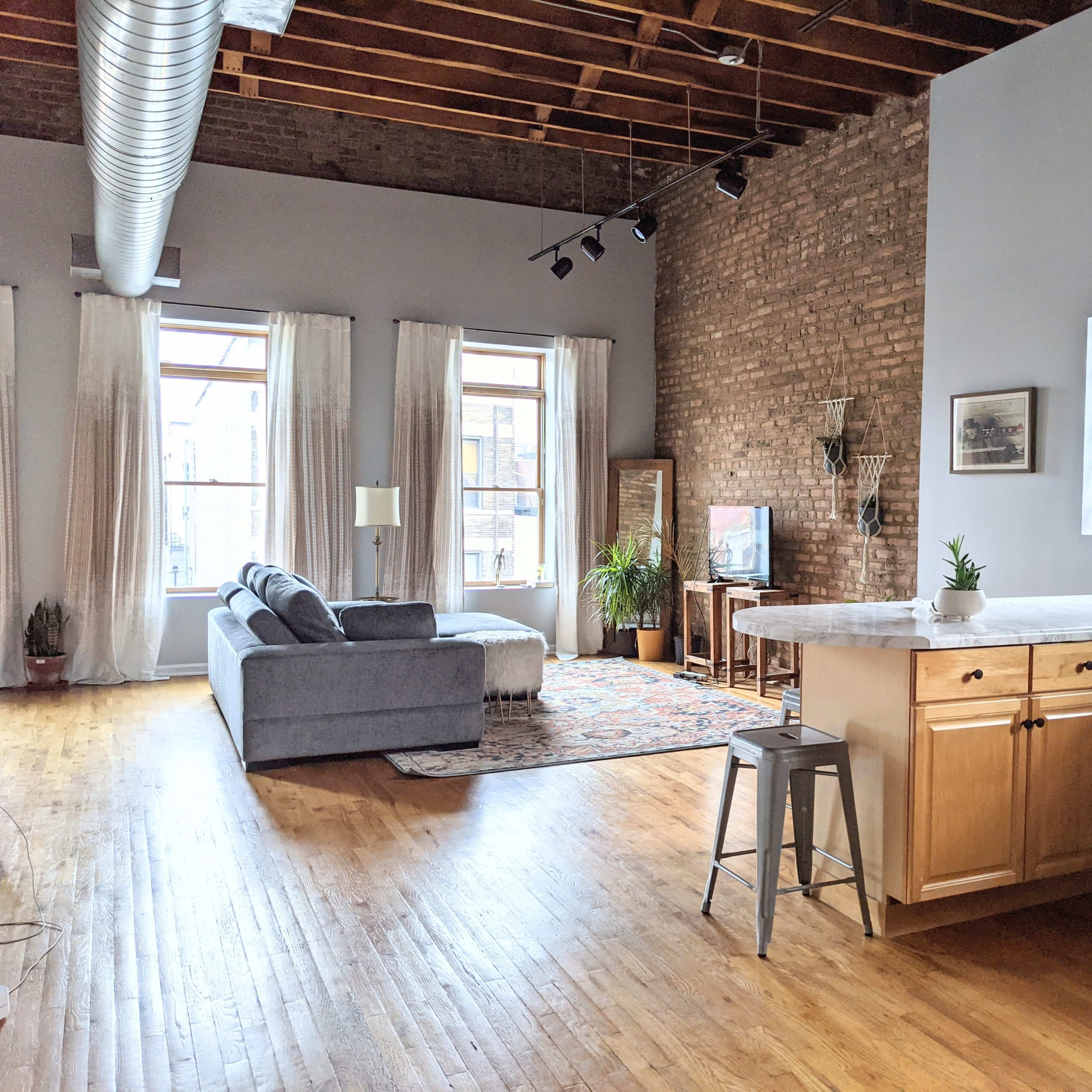 A spacious living area with a gray sofa, wooden floors, and a kitchen featuring light wood cabinets and a modern countertop.
