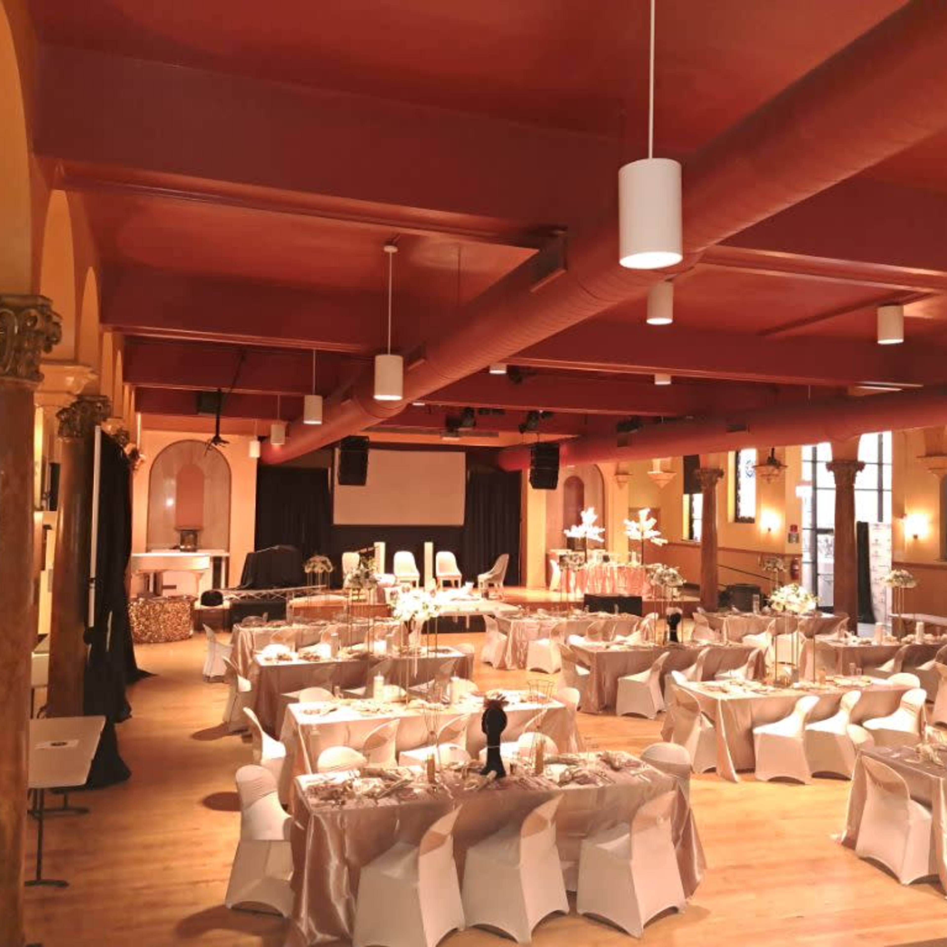 A spacious banquet hall arranged for an event, featuring elegantly set tables with white tablecloths and chairs, under warm lighting and decorative columns.