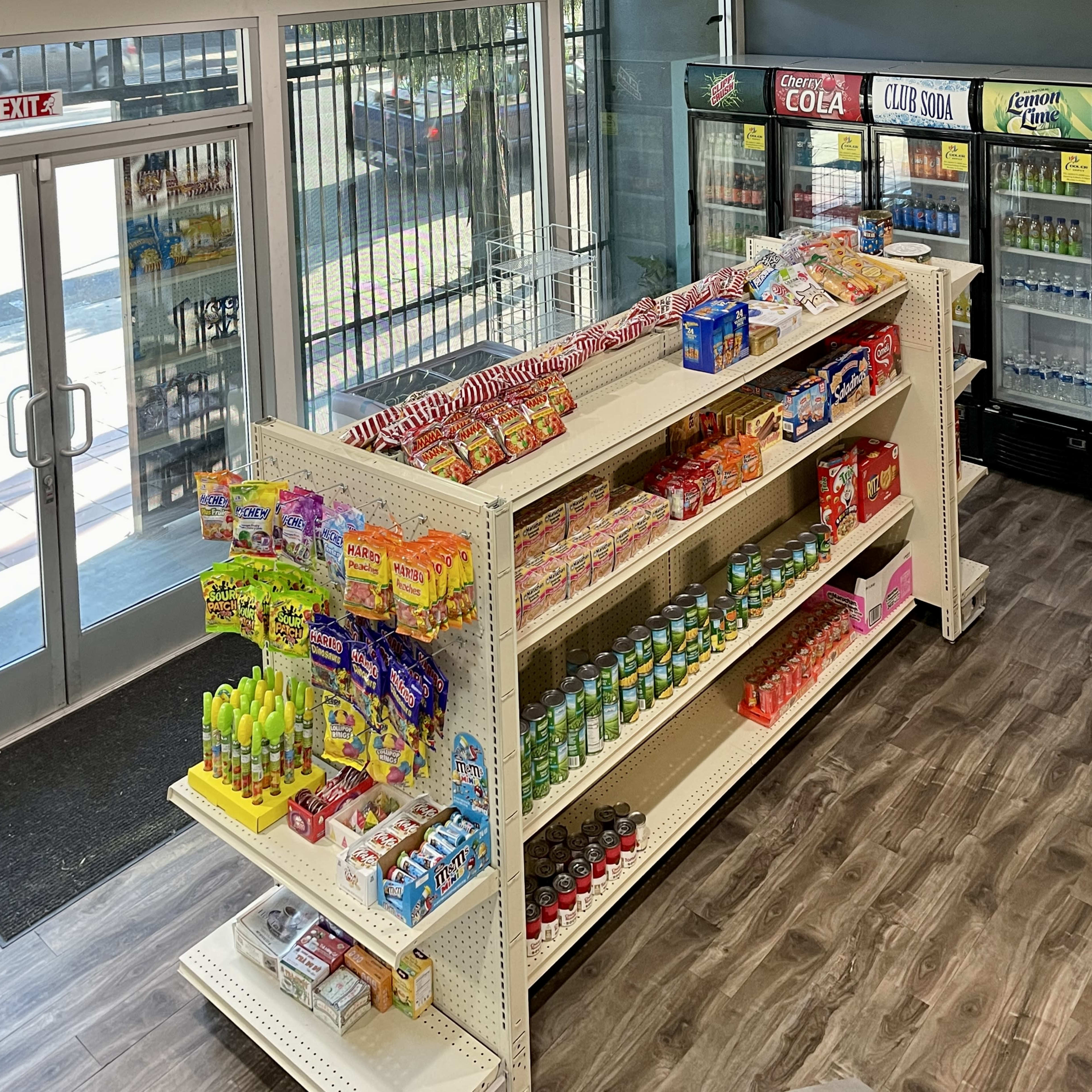 A store interior with an aisle of shelves stocked with various snacks and beverages, facing large glass doors that provide an exit.