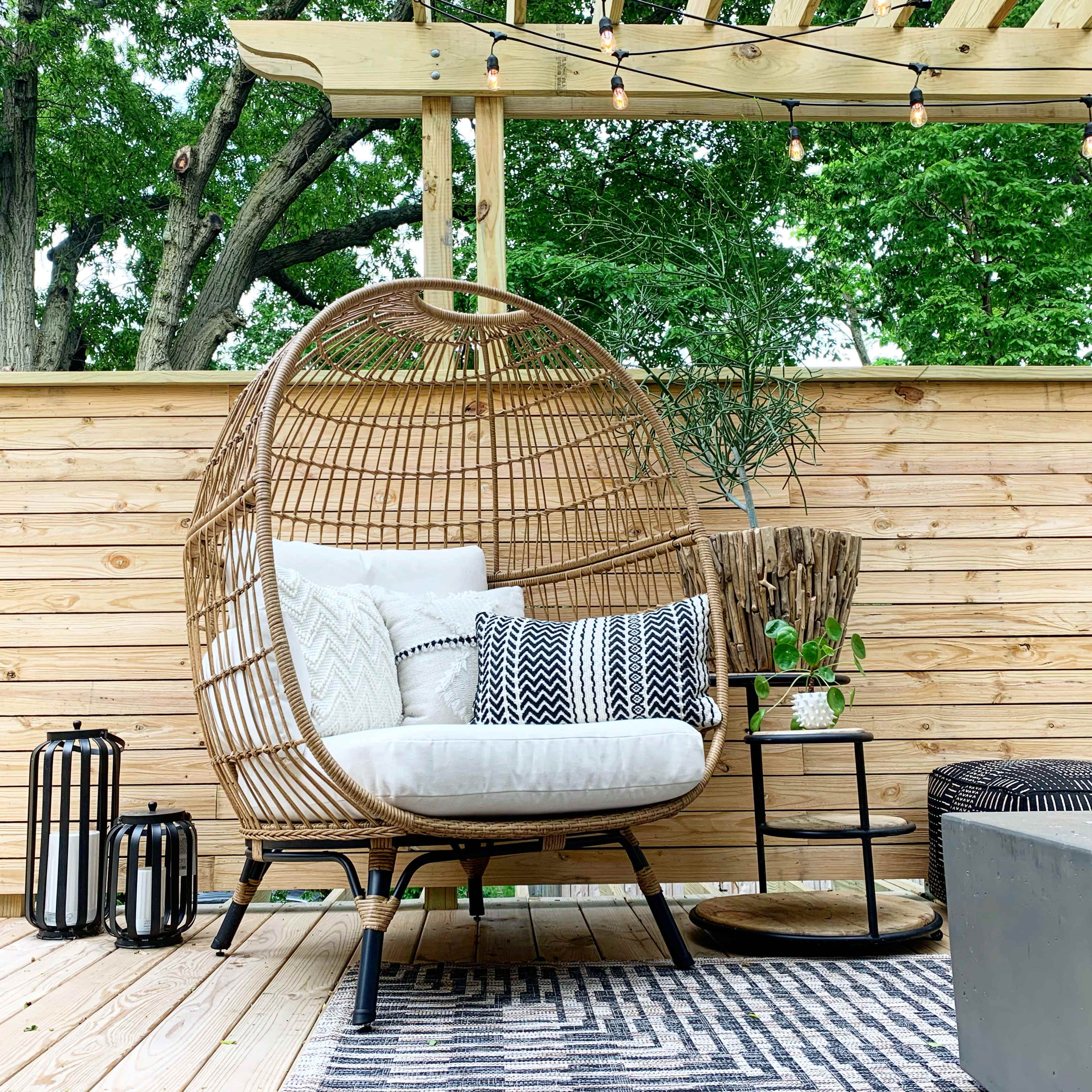A cozy wicker chair with cushions is positioned beside a small table on a wooden deck, surrounded by greenery and decorative lanterns.