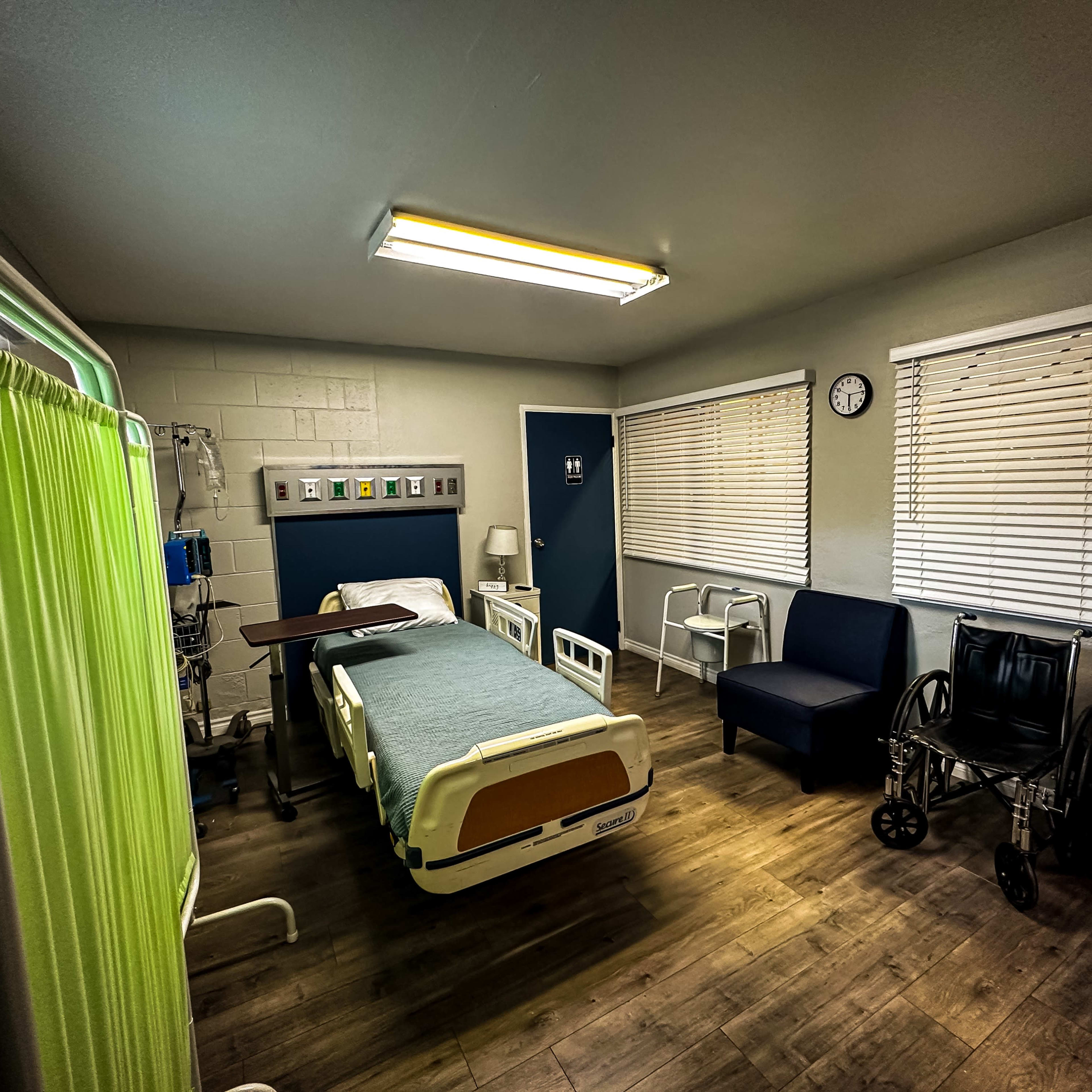 A hospital room with a bed, a wheelchair, a privacy screen, and medical equipment against the walls.