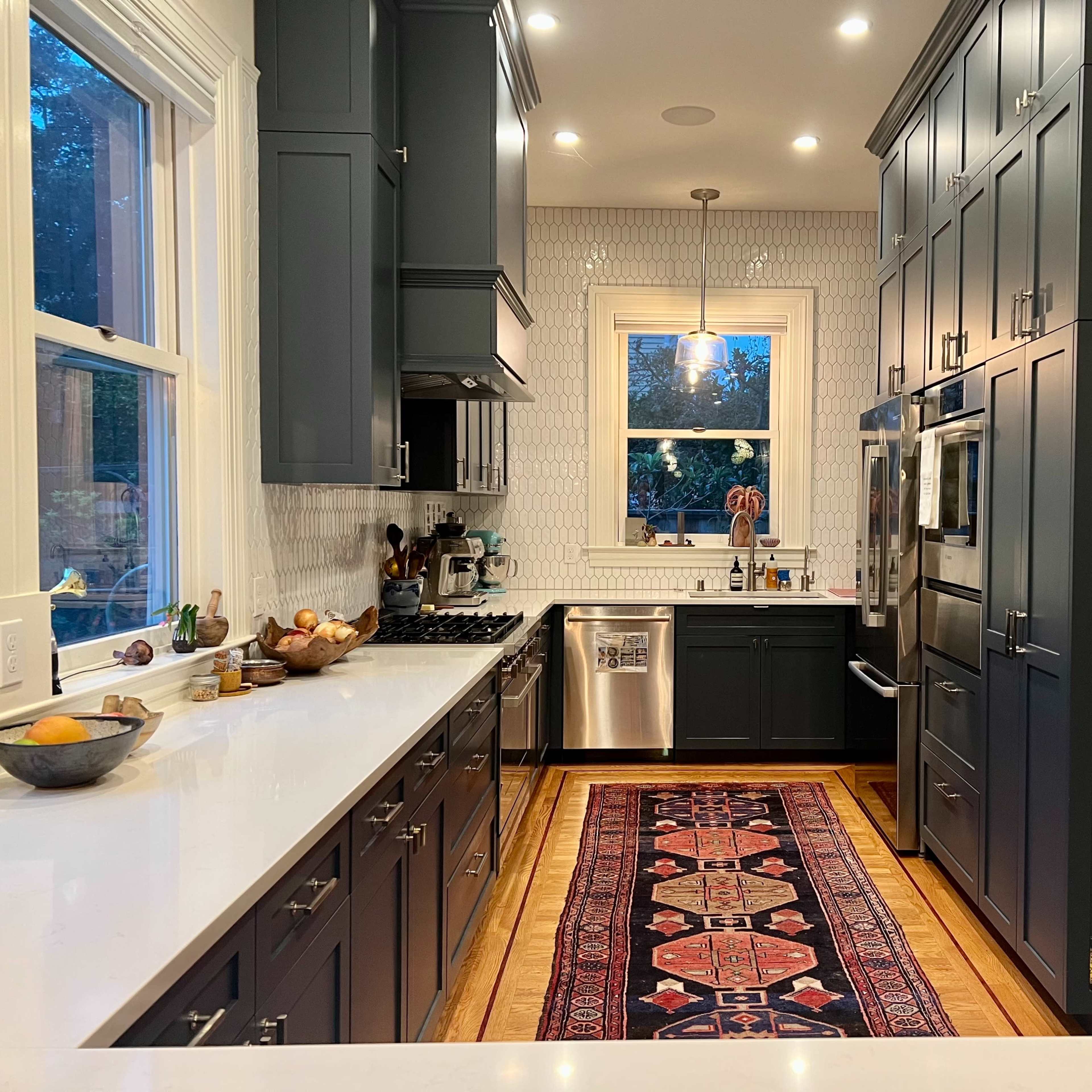 The kitchen features dark cabinets, a white backsplash, and a central island topped with a bowl of fruit, complemented by a patterned rug on the wooden floor.