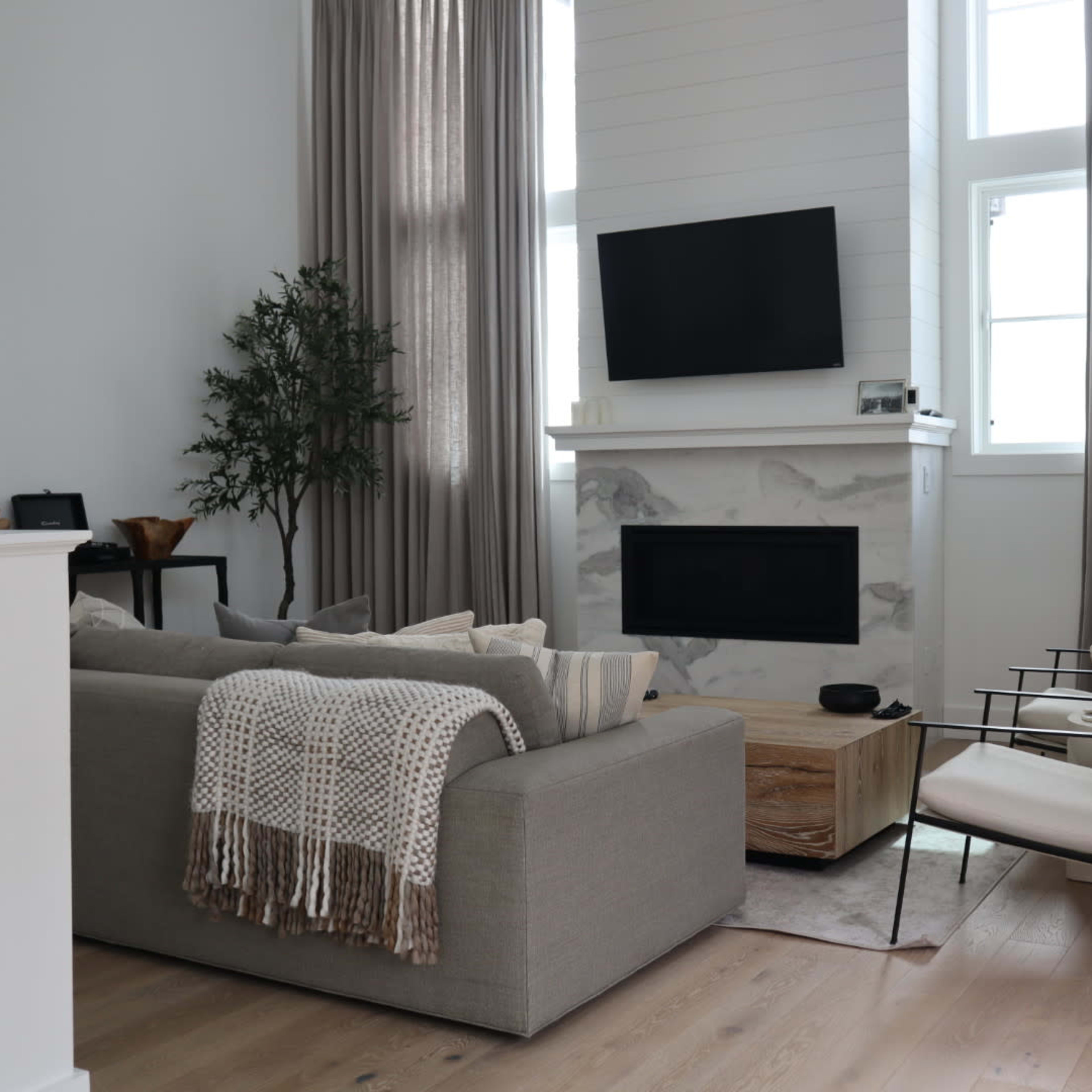 A modern living room featuring a gray sofa, a wooden coffee table, a fireplace with a white surround, and large windows covered with long, light-colored curtains.